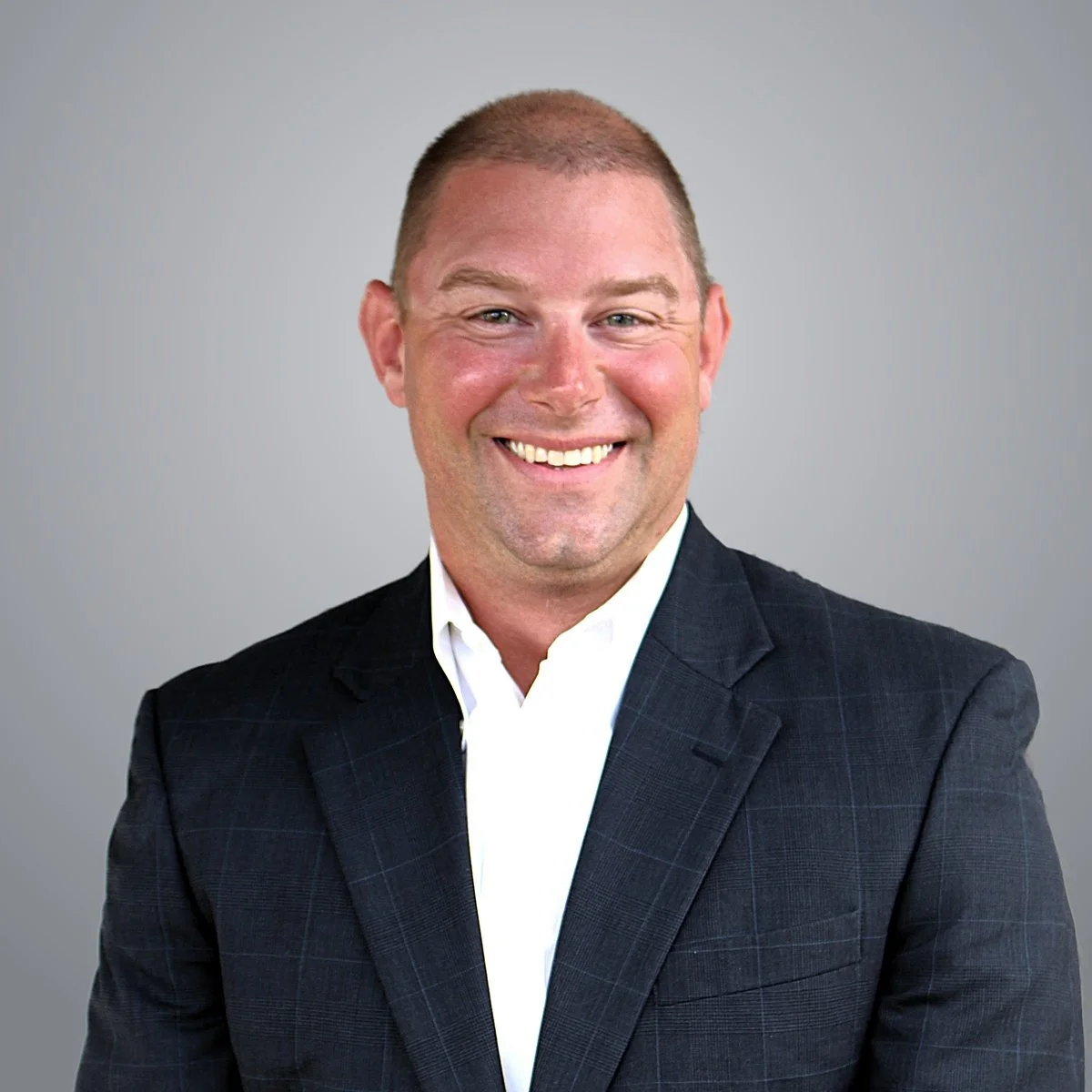 Portrait of a smiling man with short hair, dressed in a black suit and white shirt, against a gray background.