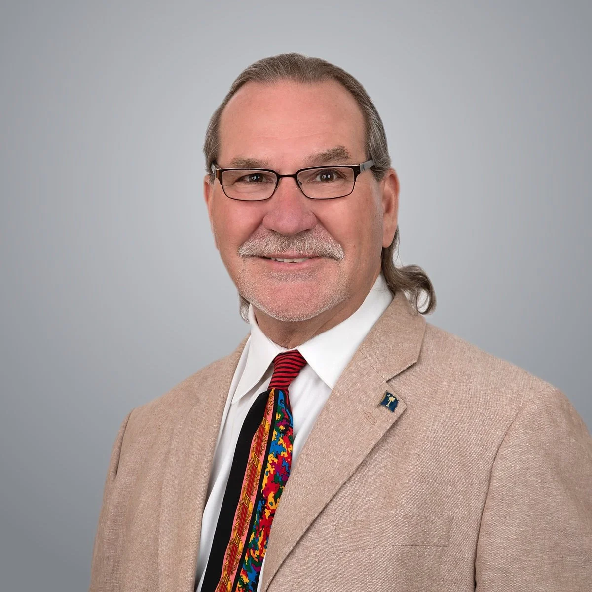 Portrait of an older man with glasses, gray hair, and a mustache, wearing a light brown blazer, a white shirt, and a colorful tie, smiling against a plain gray background.