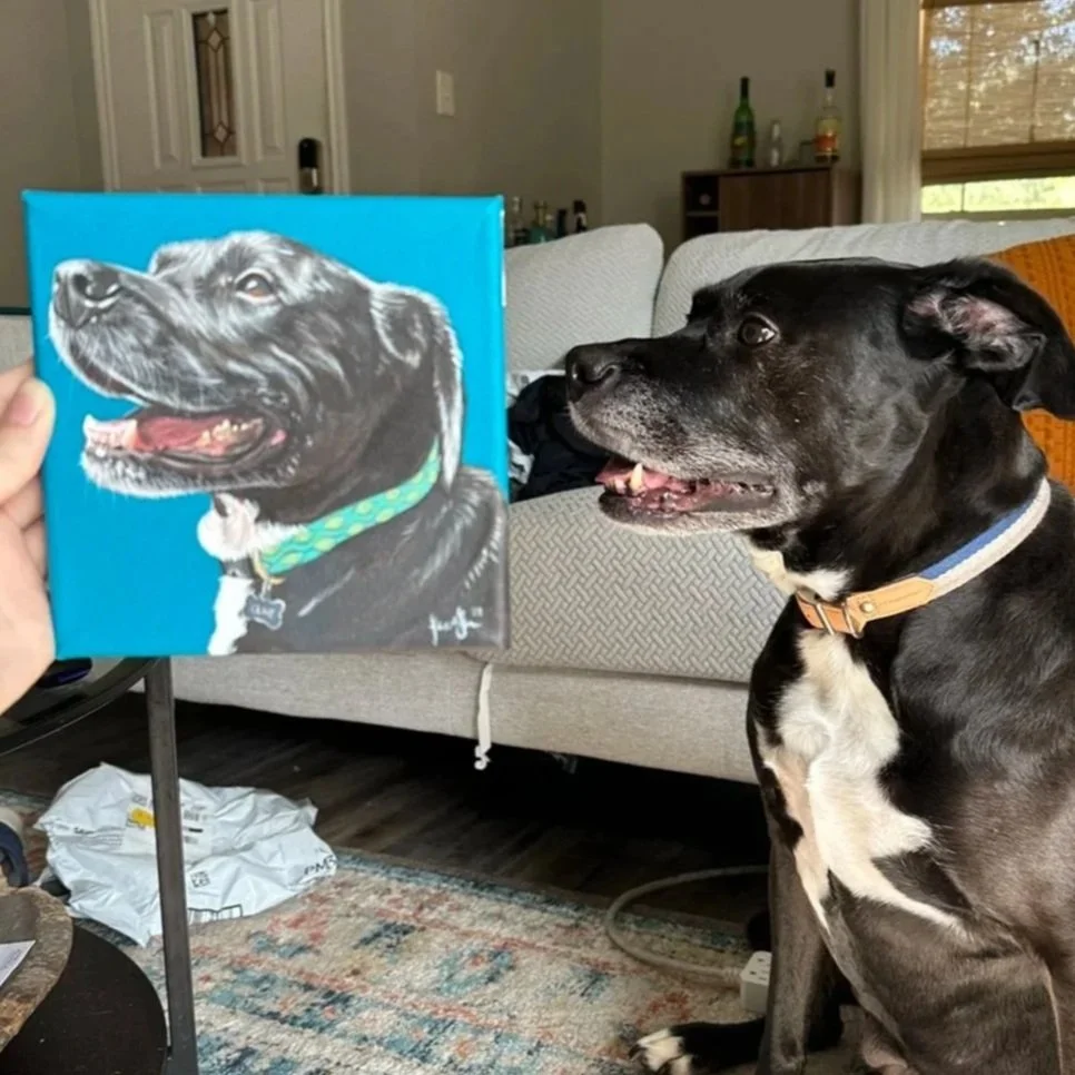 A black dog with a white chest sitting on a couch, looking at a painting of a black dog that is held up in front of it. The painting depicts the same black dog with a green collar, mouth open, and tongue out.