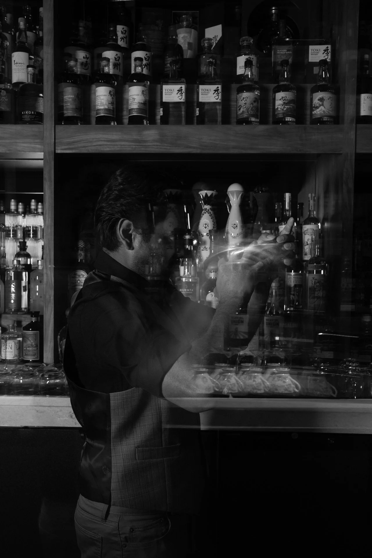 A man in a dark shirt and checked apron is taking a photo of bottles and decorative items behind a glass display case in a bar. The background shows shelves filled with liquor bottles.