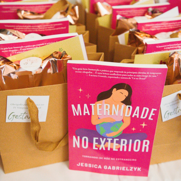 A copy of the book Maternidade no Exterior by Jessica Gabrielczyk displayed in front of several brown paper gift bags filled with event goodies.