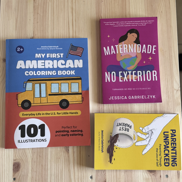 Flat lay of publications by author Jessica Gabrielzyk, including My First American Coloring Book, Maternidade no Exterior, and Parenting Unpacked: Parenting Through The Loss of Self, displayed on a light wooden surface.