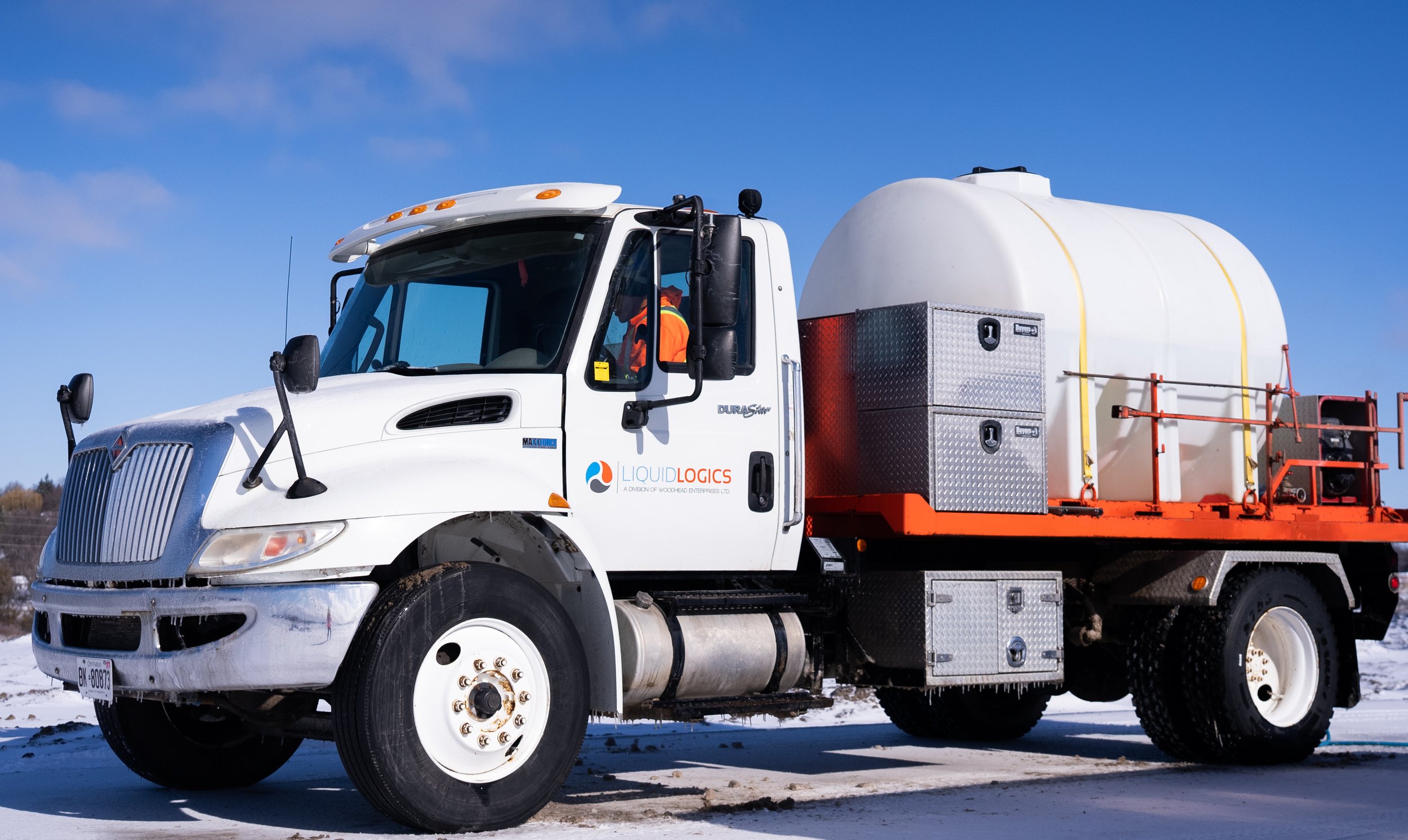 Large white truck parked on snowy ground, carrying a cylindrical tank on its bed with "Liquid Logics" branding, under a clear blue sky.