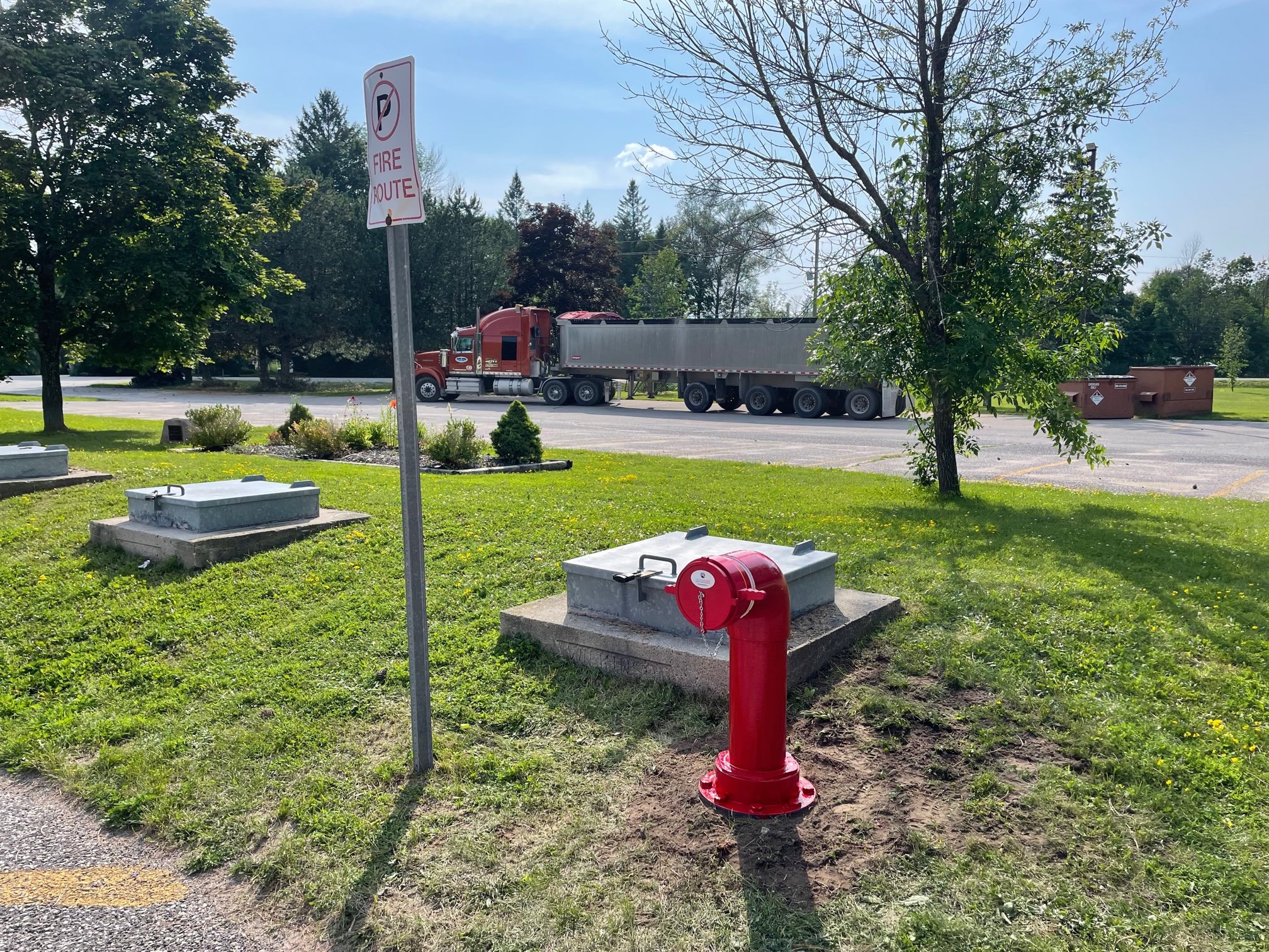 Red fire hydrant on grass near "No Parking Fire Route" sign, adjacent to large truck in parking lot; surrounded by greenery.