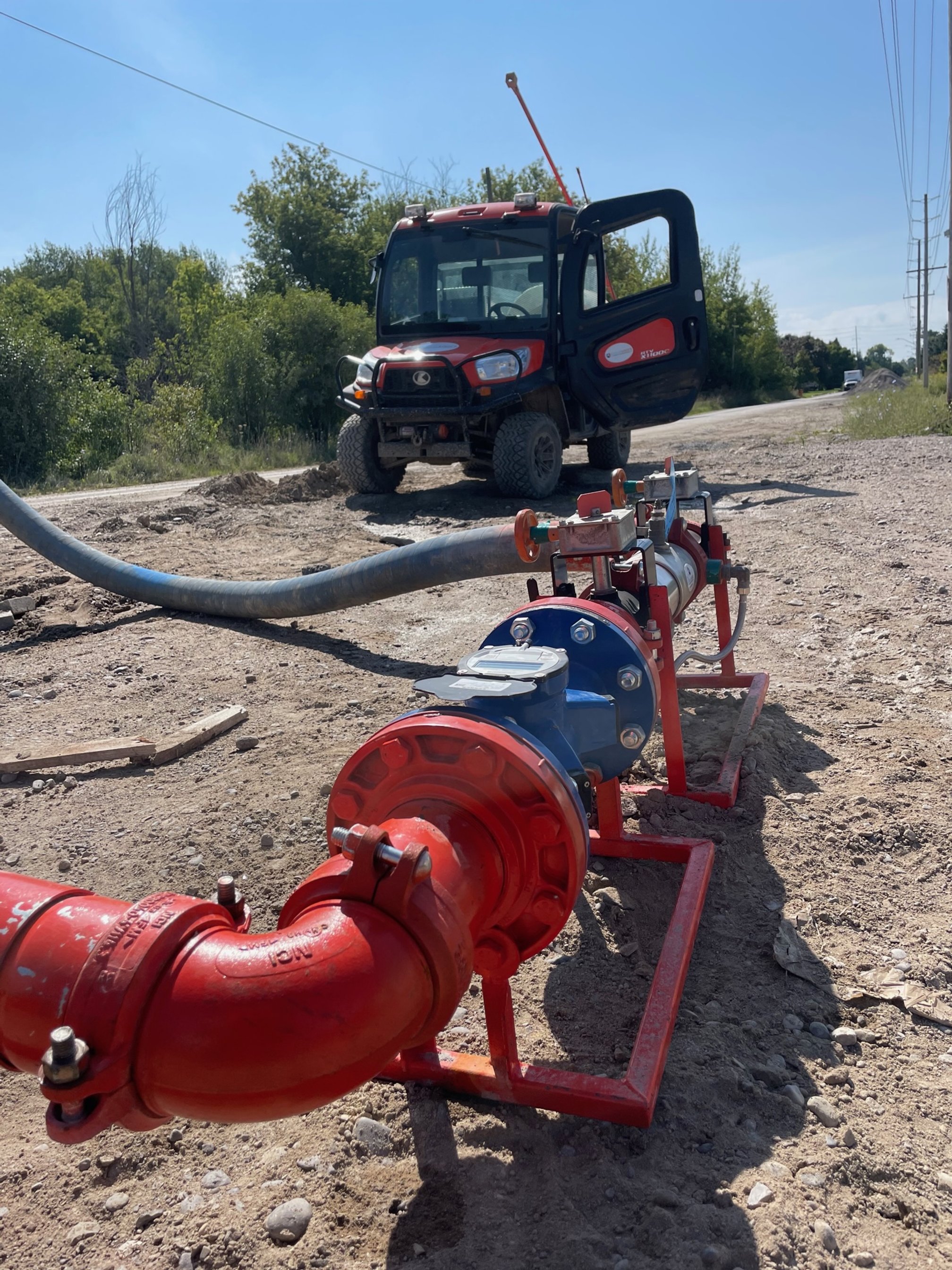 A red utility vehicle parked on a dirt road with piping equipment and a hose in the foreground, surrounded by trees.