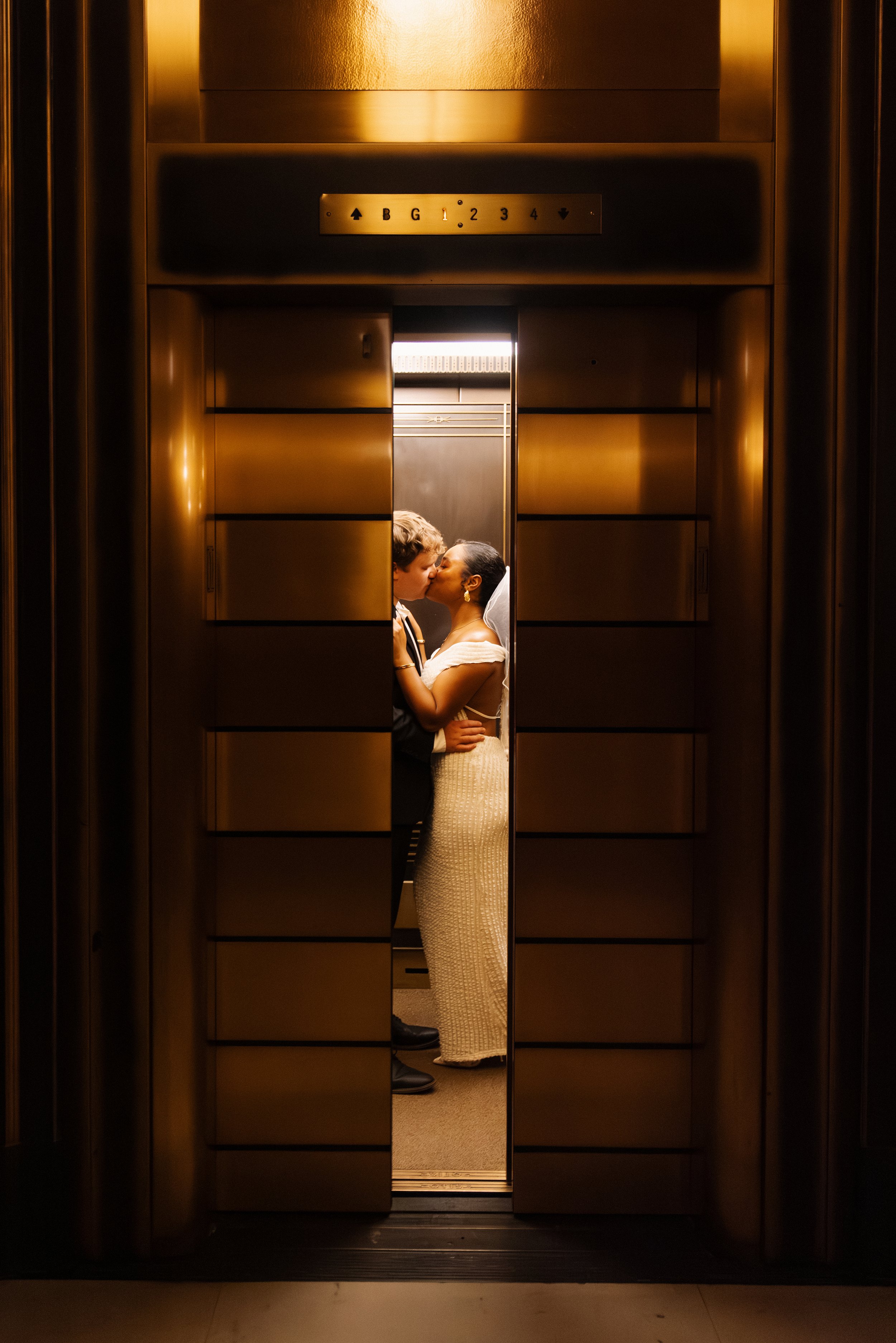 A bride and groom kissing inside an elevator with reflective bronze interior walls.