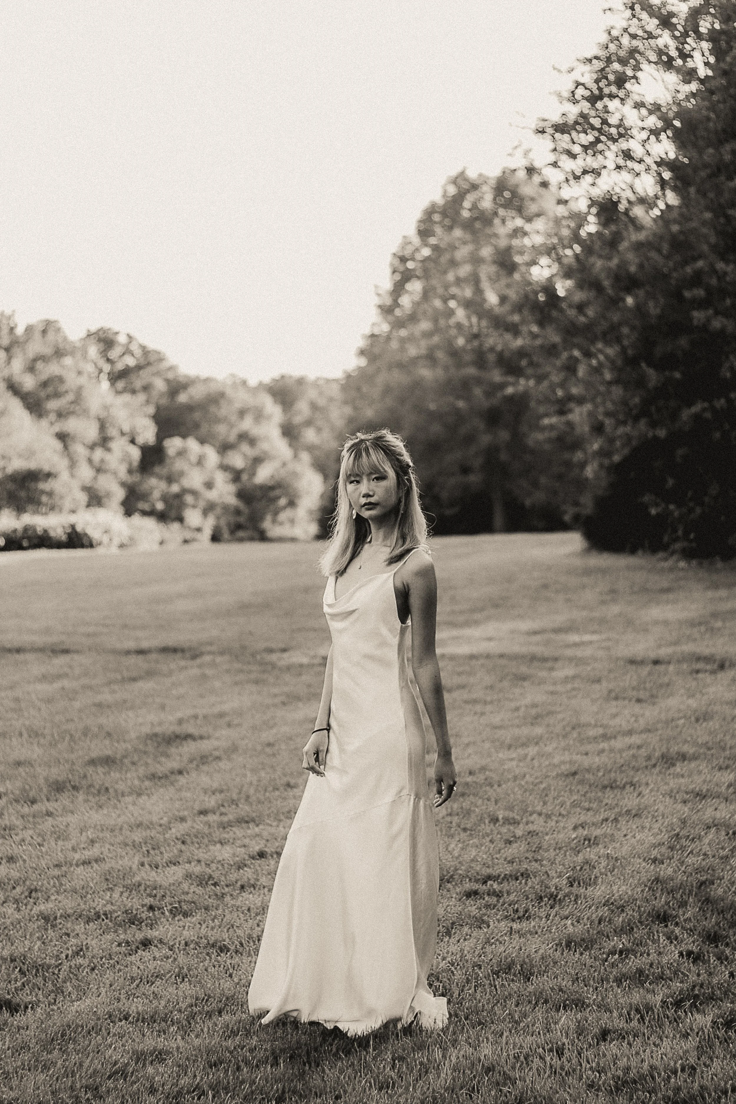 A woman in a long, elegant dress standing on a grassy field with trees in the background, in black and white.