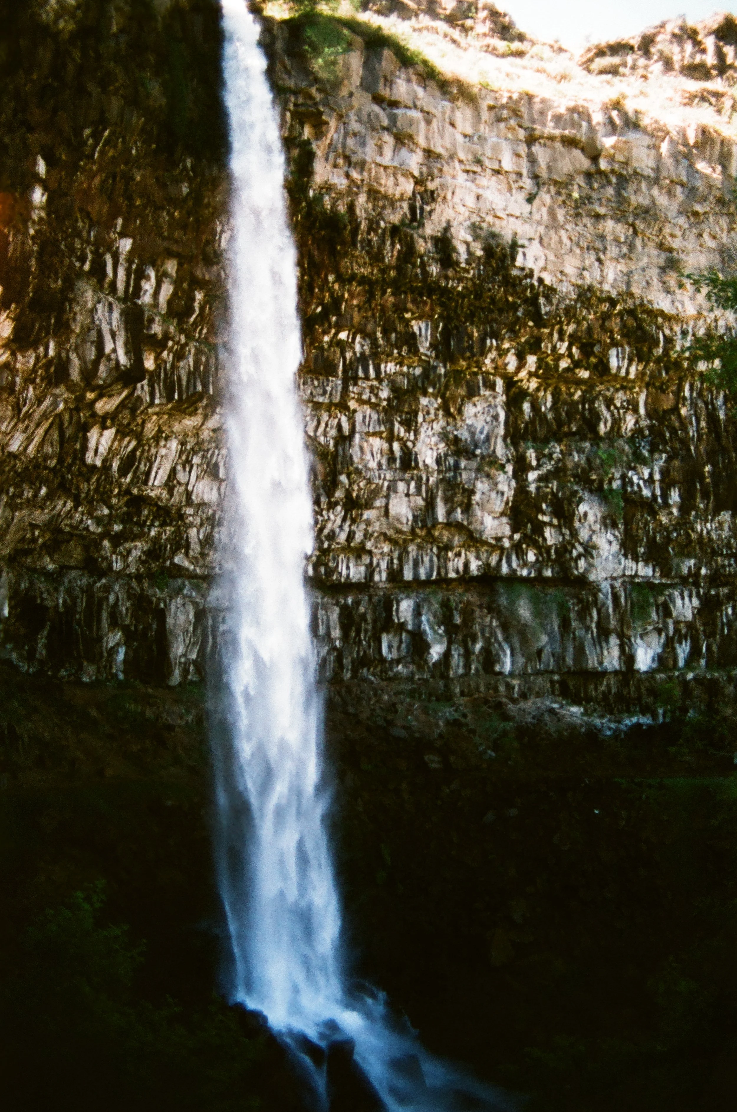 A tall waterfall cascading down a rocky cliffside into a dark pool below, surrounded by greenery.