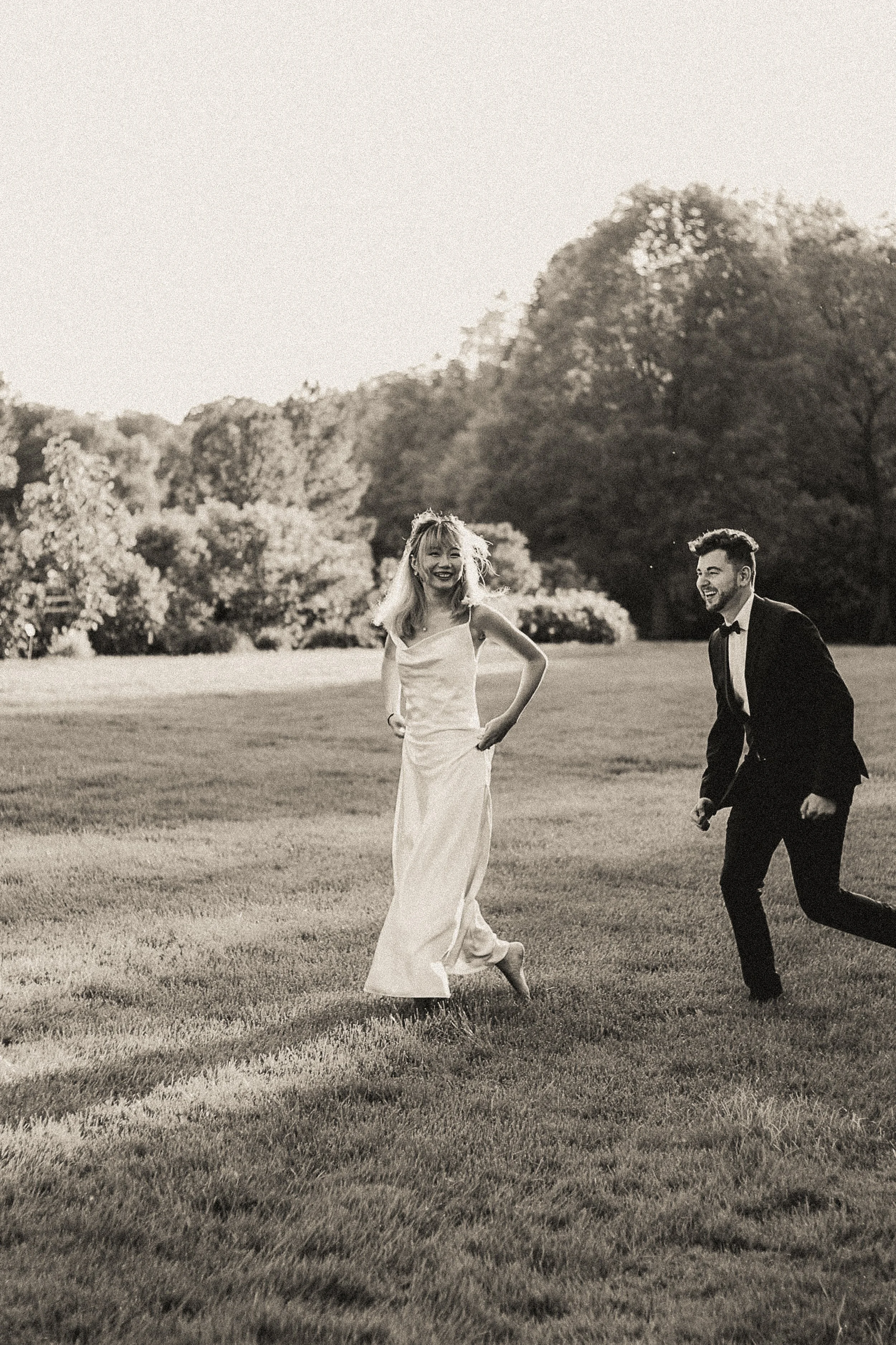 Black and white photo of a man in a tuxedo and a woman in a wedding dress running and laughing on a grassy field with trees in the background.