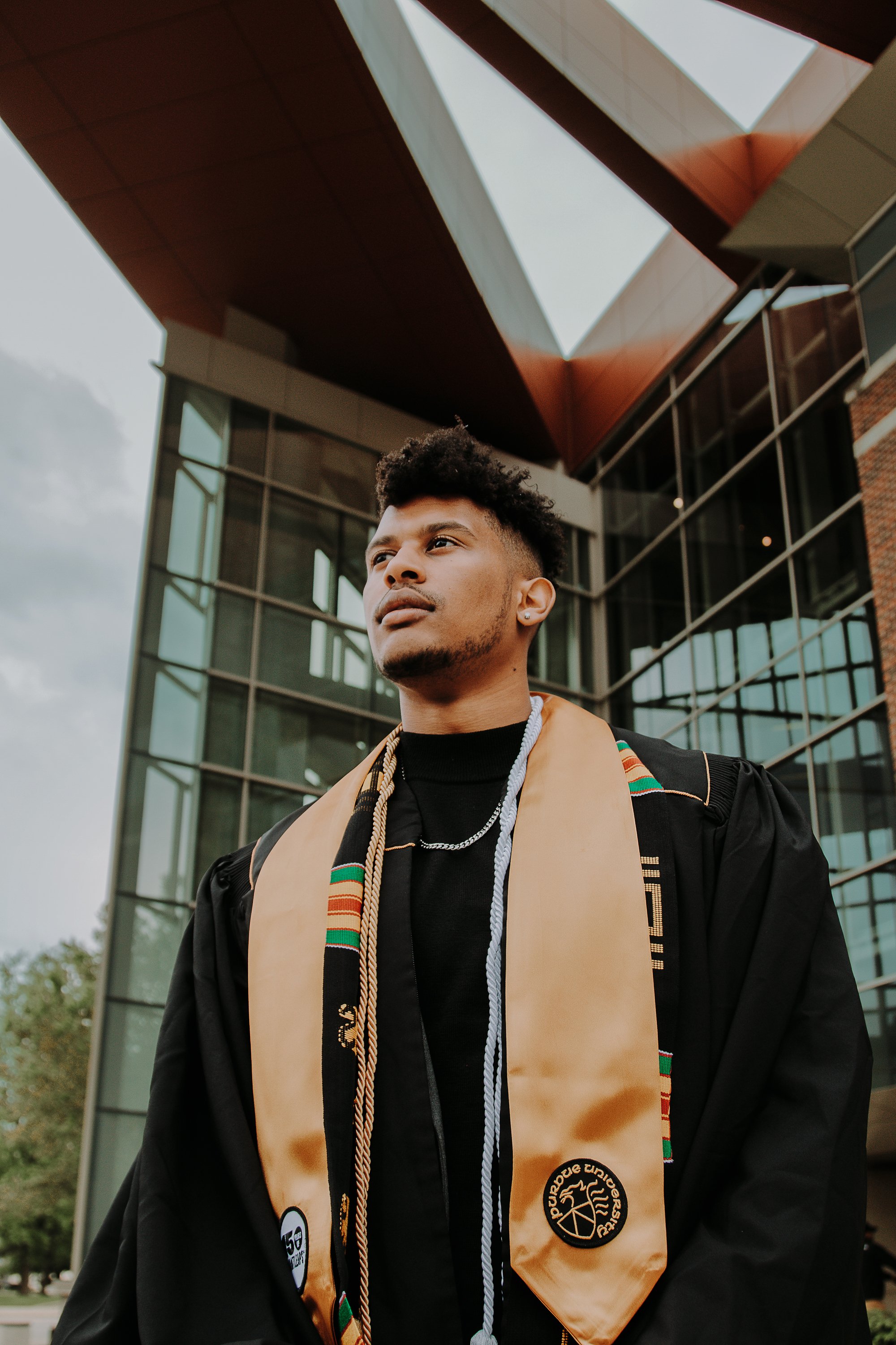 Young man in graduation gown and honor cords standing outside modern glass building.
