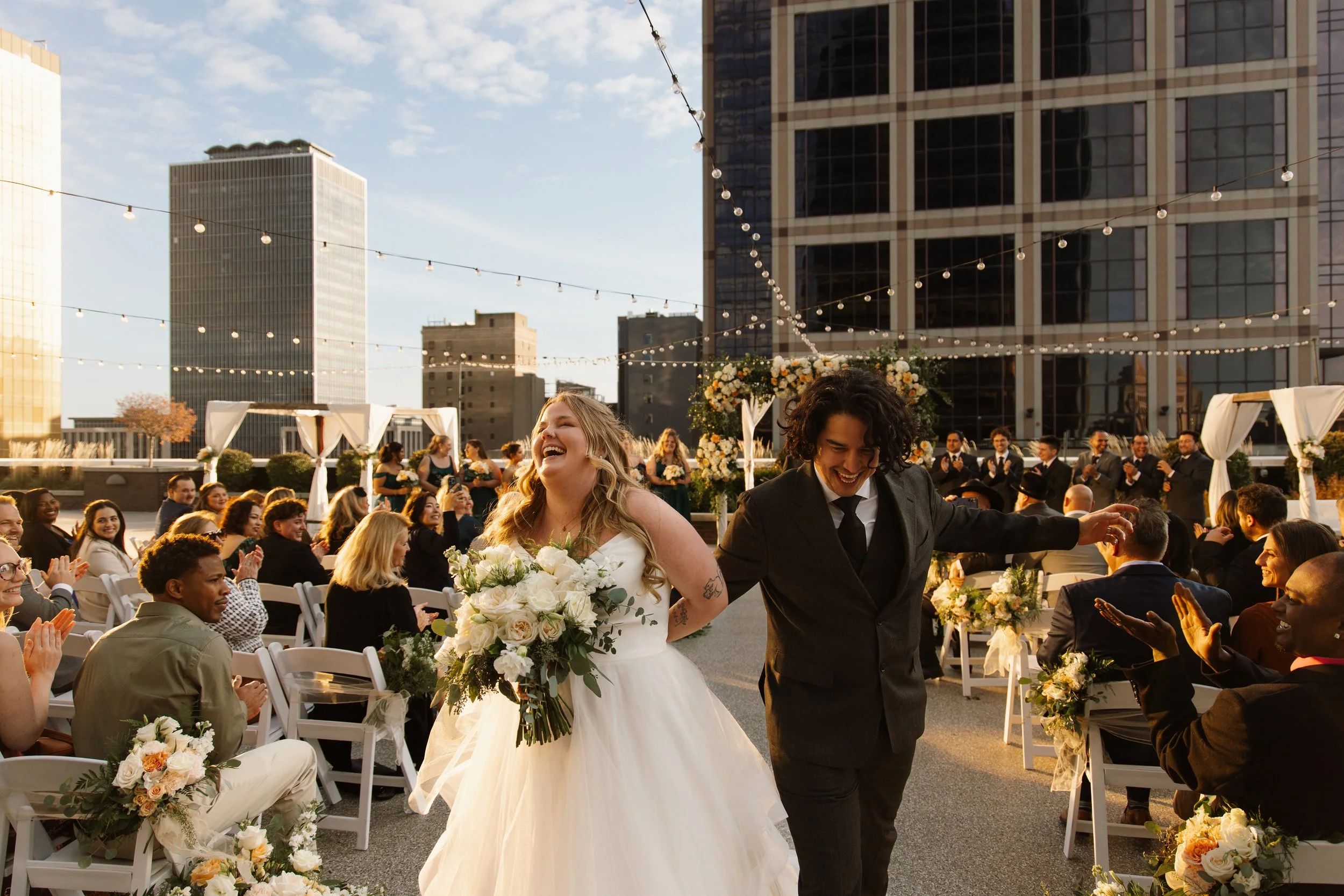 A newly married couple, a woman in a white wedding dress and a man in a dark suit, walking down the aisle at an outdoor wedding ceremony on a rooftop during sunset. Guests are seated and clapping, and string lights and floral decorations surround the scene.