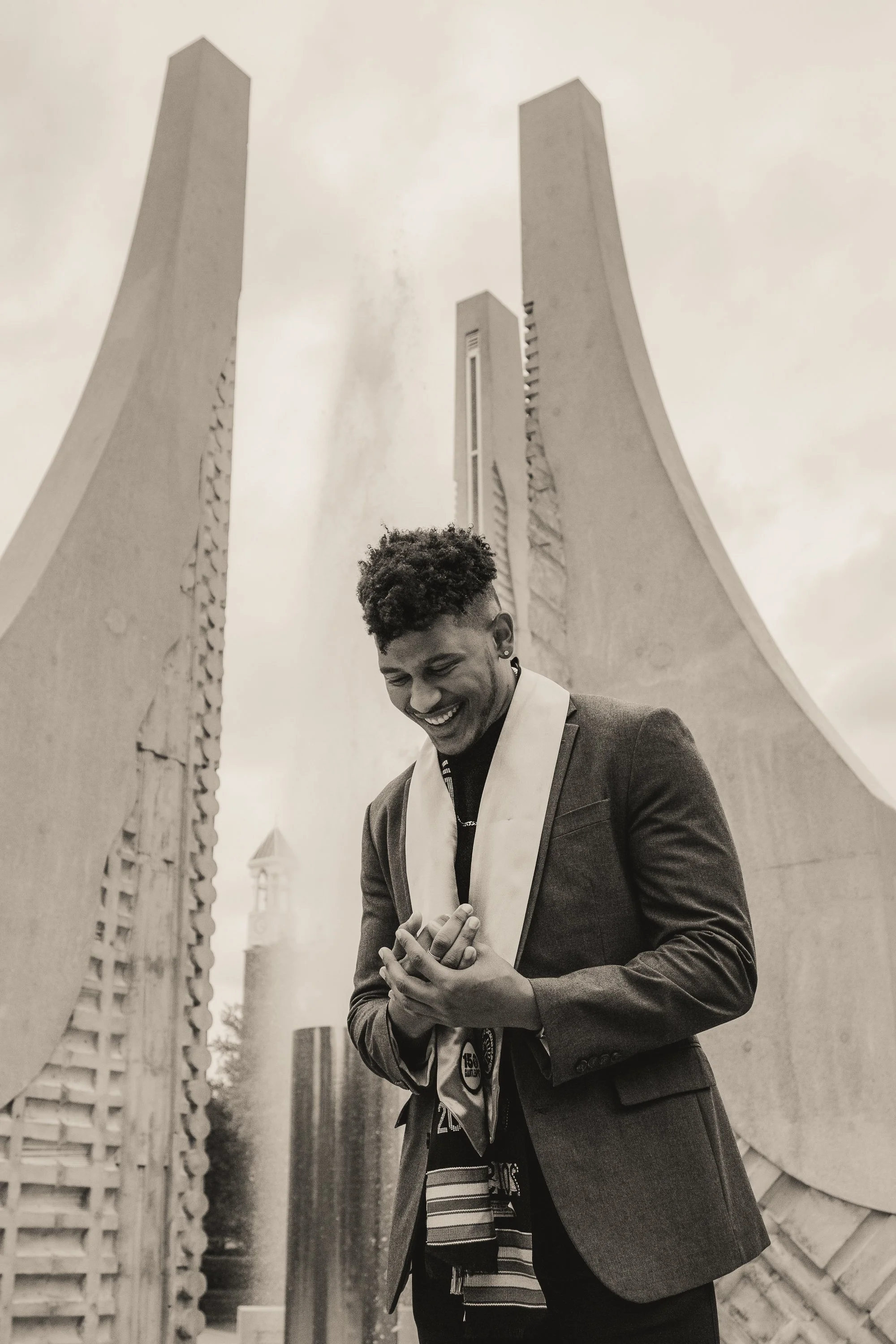 Black and white photo of a young man in a tuxedo and scarf smiling with hands clasped, standing outdoors in front of a modern architectural structure.
