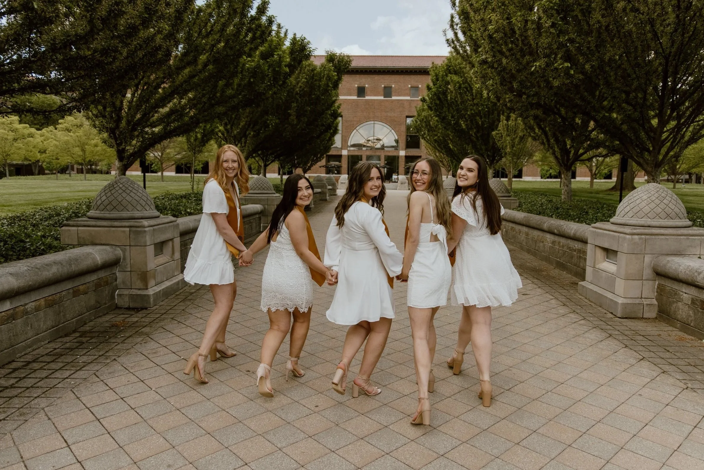 A group of six women standing outdoors, holding hands, wearing white dresses and beige high heels, smiling, with a brick building and trees in the background.