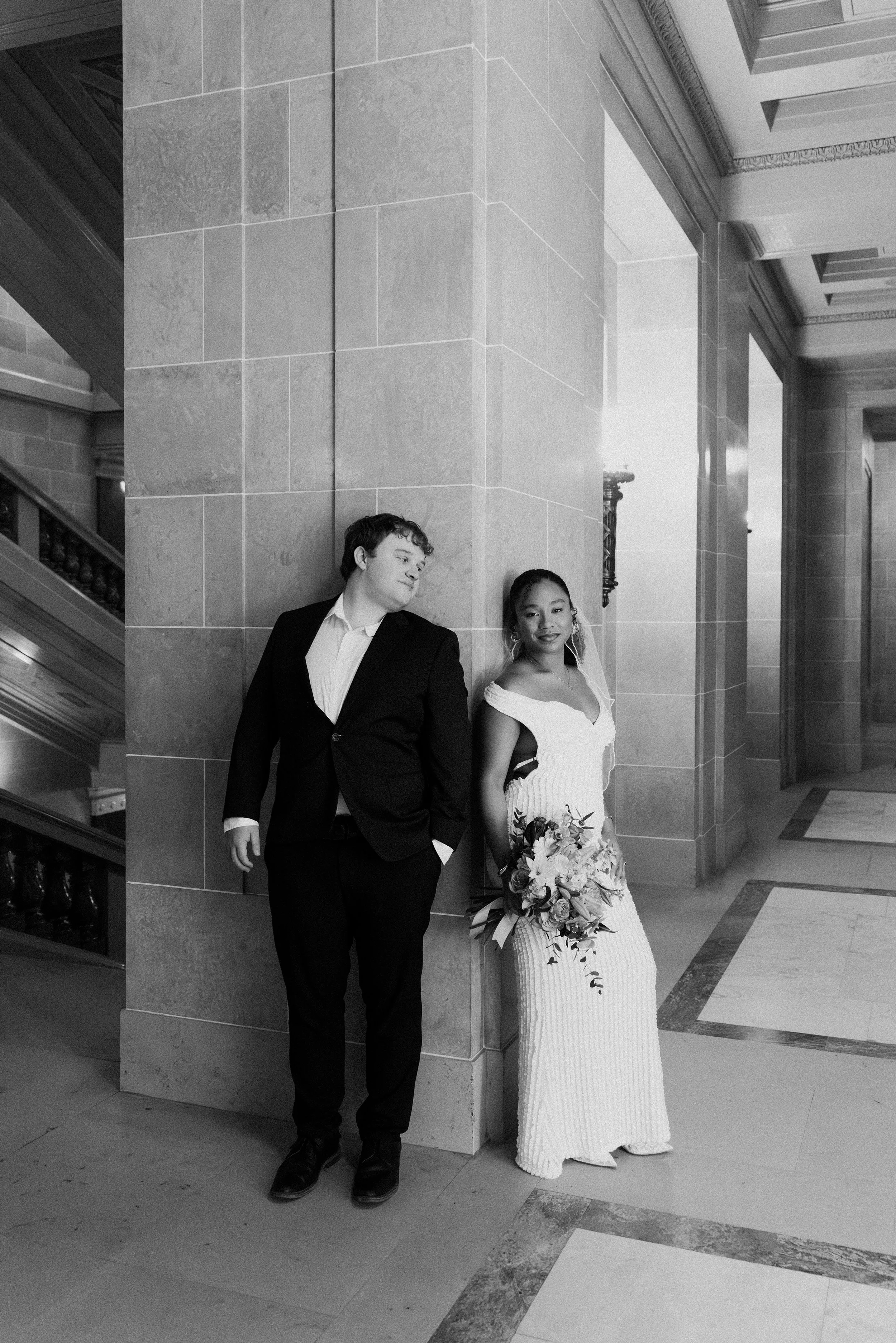 A black and white photo of a couple in wedding attire standing against a stone column in a grand interior building. The man is in a suit with hands in his pockets, looking at the woman. The woman is in a long dress, holding a bouquet, looking at the 