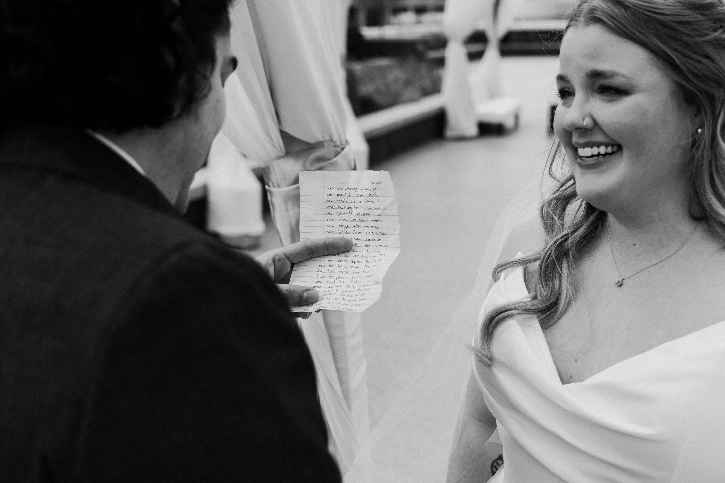 A man exchanging wedding vows with woman in a wedding dress, with the man holding a handwritten note and the woman smiling with visible wedding jewelry and veil.