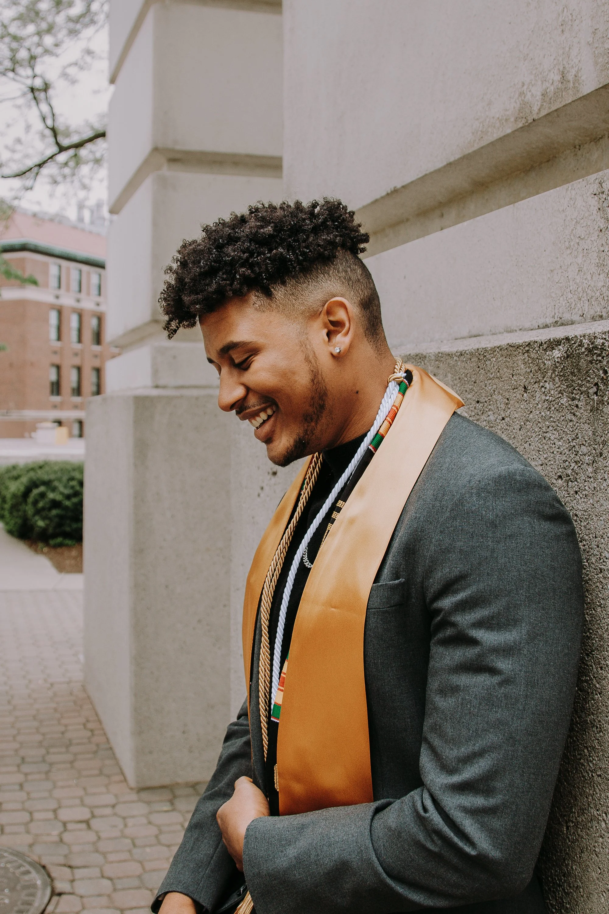 A young man with curly hair and a beard smiling while leaning against a concrete wall outdoors, wearing a dark suit and a colorful stole.