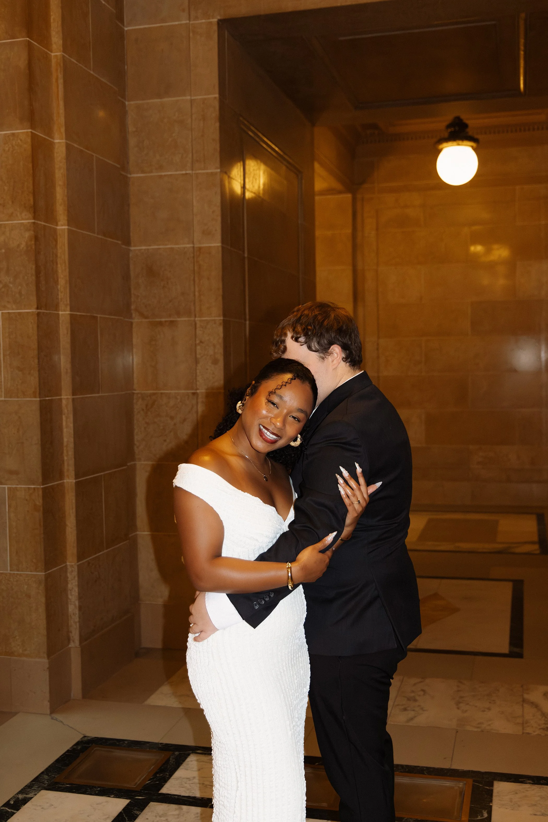 A happy couple embraces in a warmly lit indoor setting, with the woman wearing a white off-the-shoulder dress and the man in a black suit.