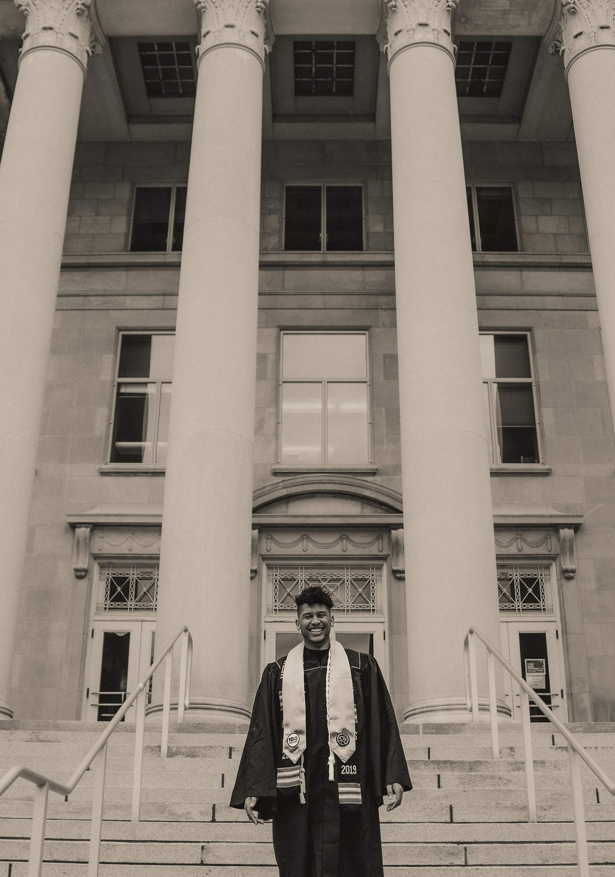A smiling young man in graduation regalia standing on the steps of a large, classical-style building with tall columns.