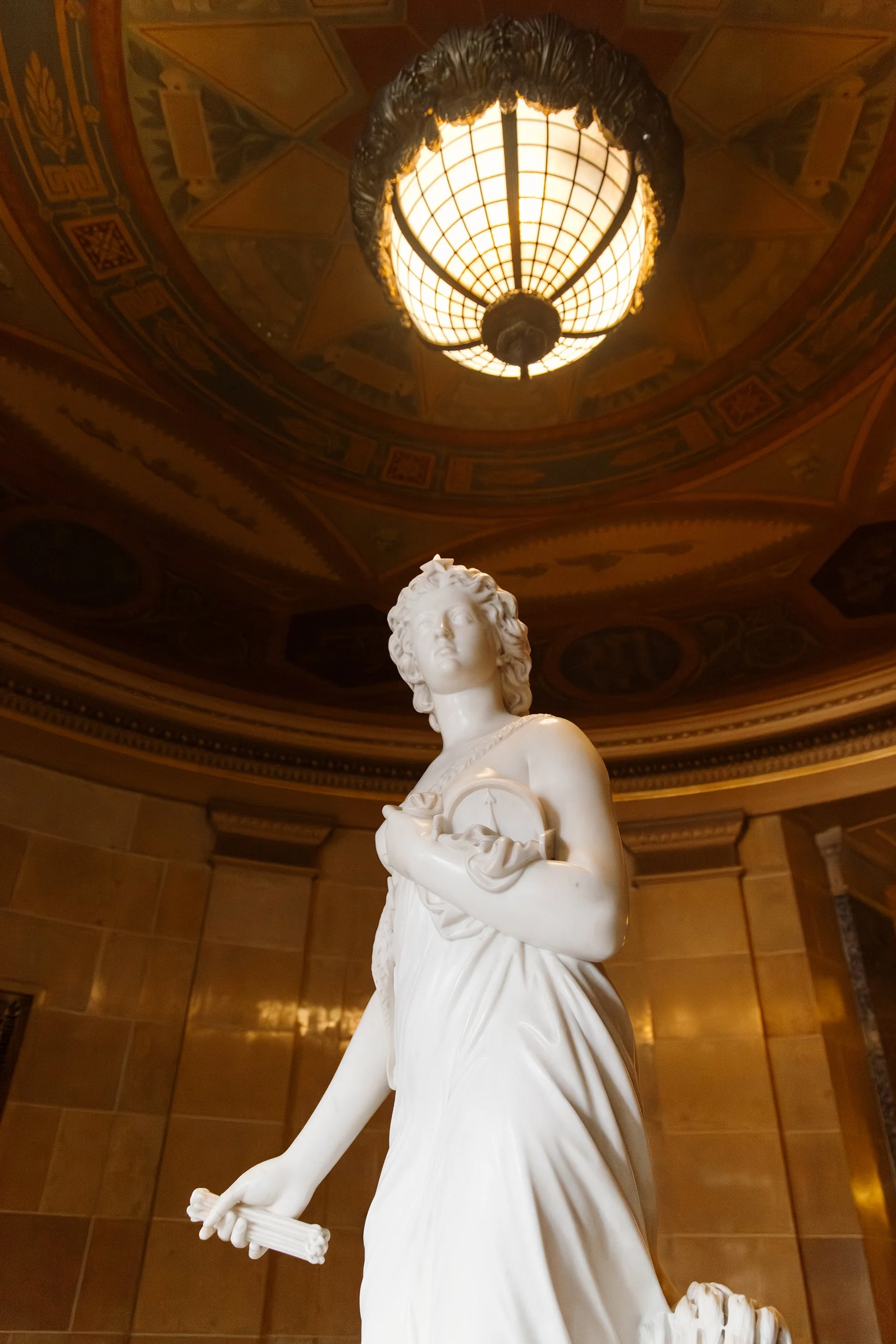 A white marble statue of a woman holding a scroll in her right hand and a round object near her chest, standing in an ornately decorated room with a large, lit ceiling light fixture.