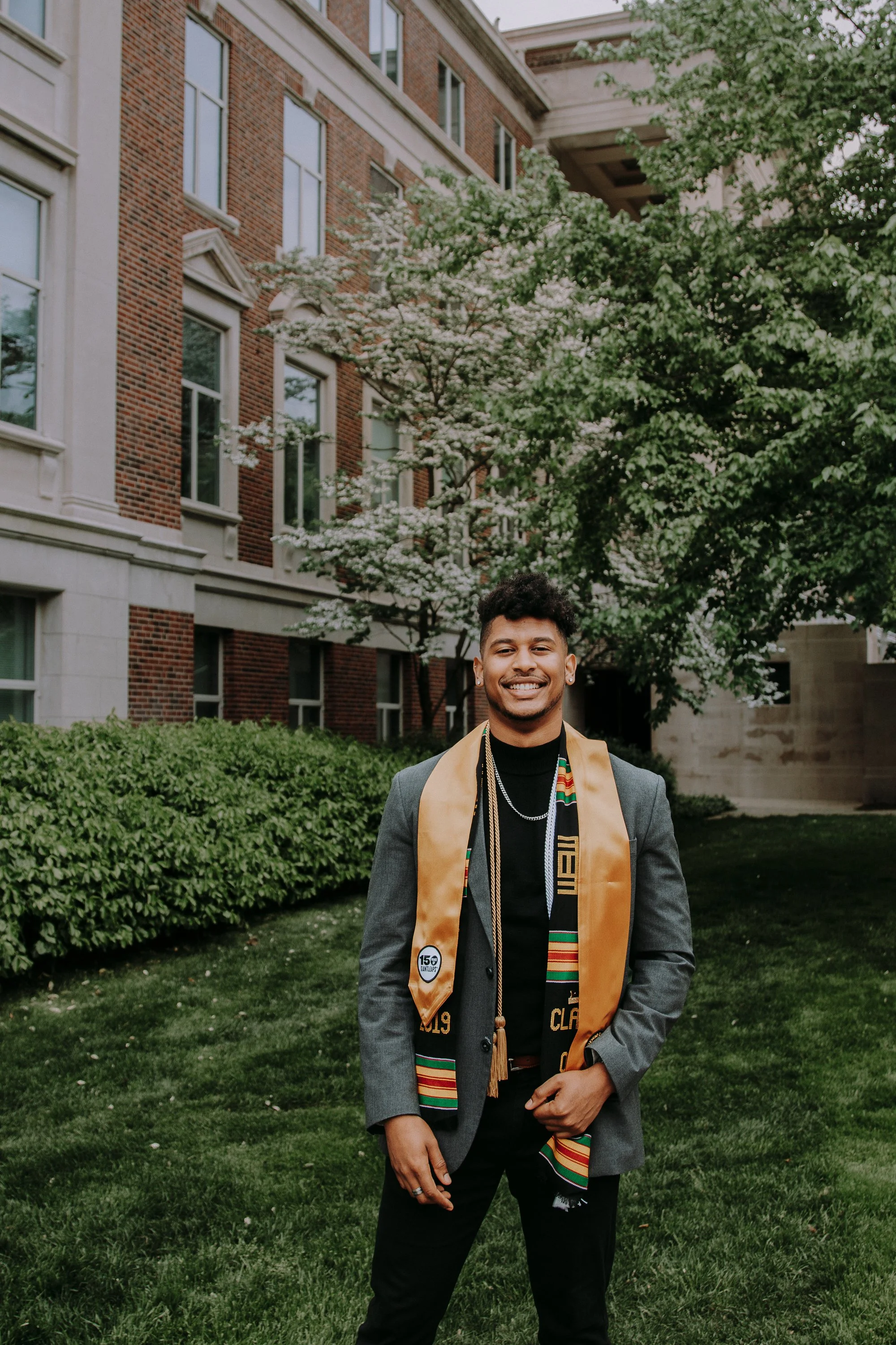 A young man in a graduation gown and stole standing outdoors on a grassy area with trees and a brick building in the background, smiling at the camera.