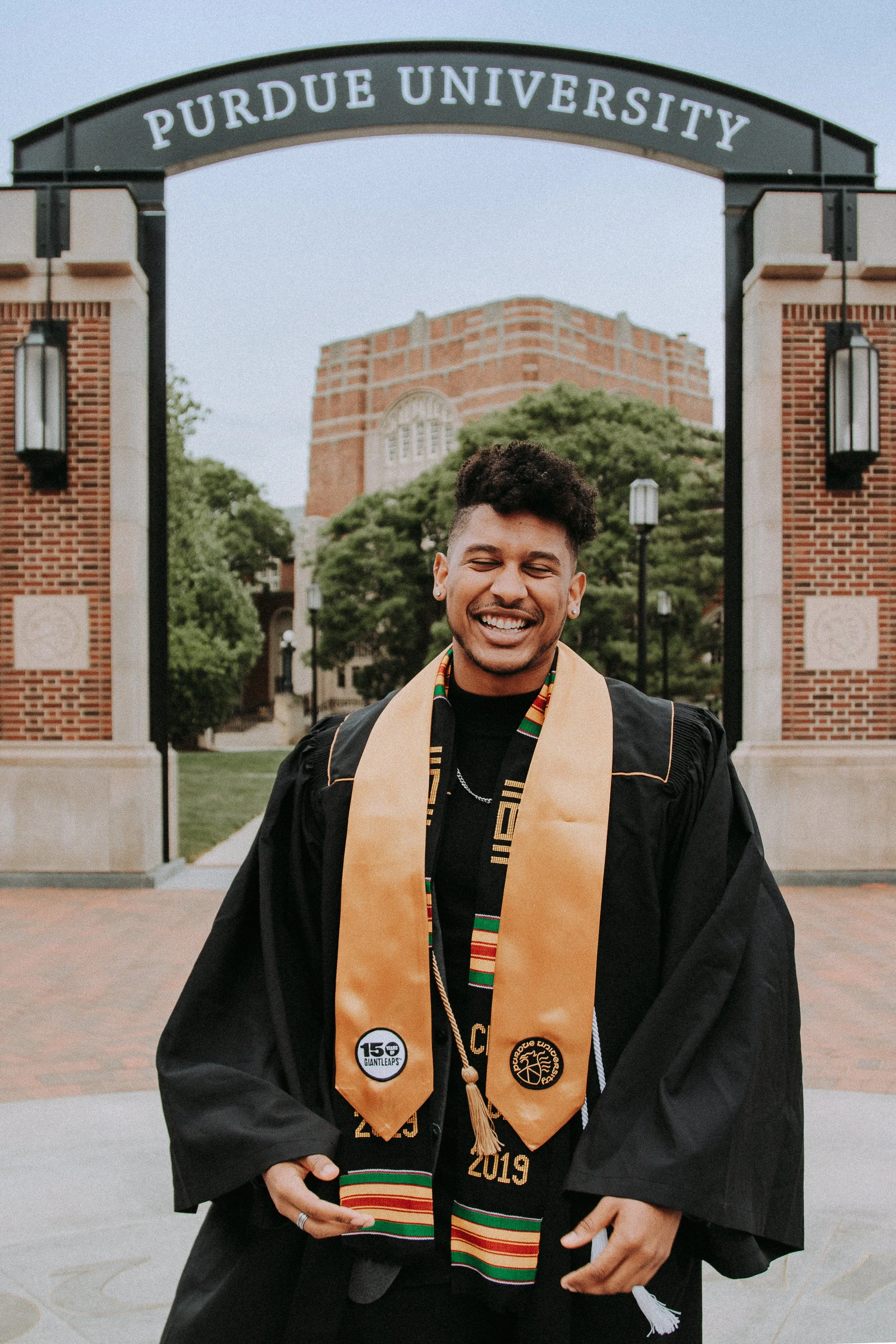Young man in graduation gown and cap standing in front of Purdue University archway, smiling with a gown sash and cap tassel.
