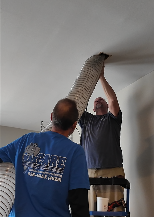 Man using a red vacuum hose to remove debris from ceiling vent in a bright living room.