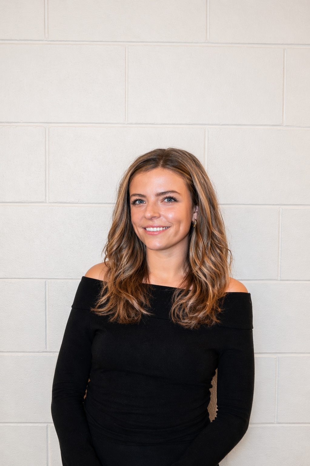 A woman with light brown, wavy hair and blue eyes, smiling, wearing a black off-the-shoulder top, standing against a plain white brick wall.