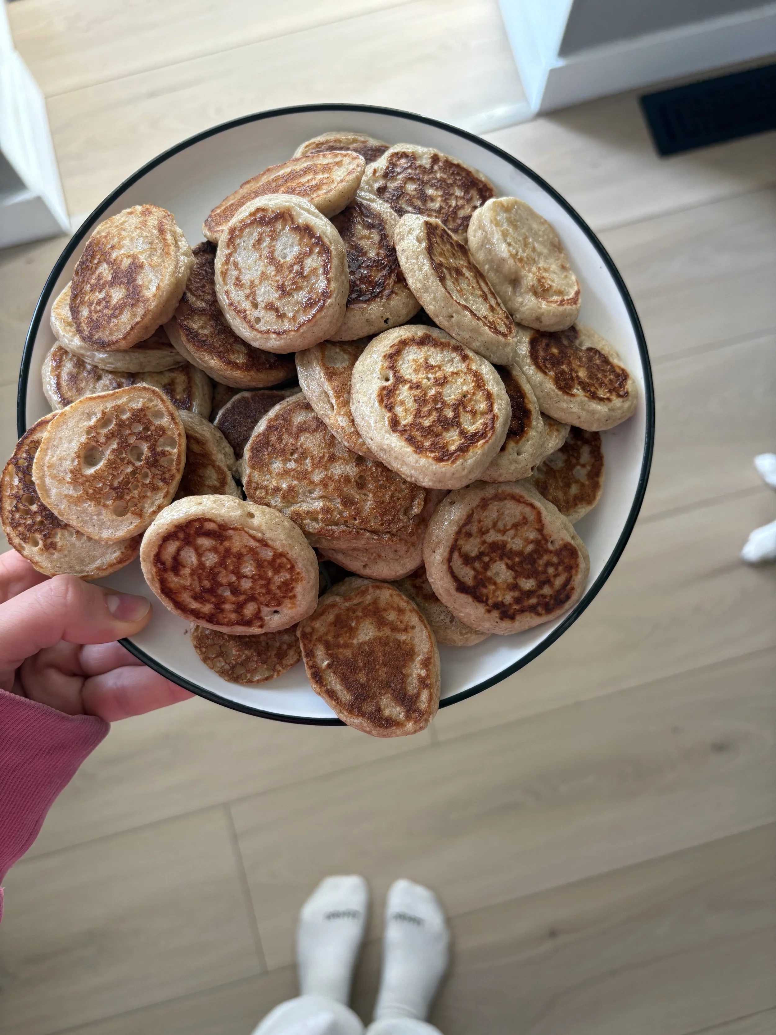 A bowl of cooked, golden-brown mini pancakes being held by a person in a pink sweatshirt. The person is standing on a light wooden floor, and their striped socked feet are visible in the foreground.