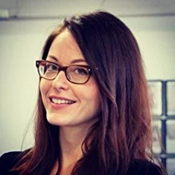 A woman with long dark hair and glasses smiling at the camera in an indoor setting.
