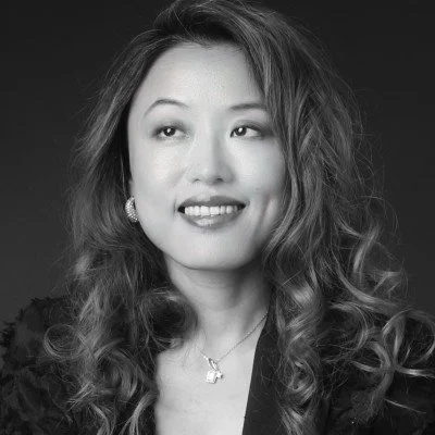 A Black and white portrait of an Asian woman with wavy hair, wearing earrings and a necklace, looking to her right with a slight smile.