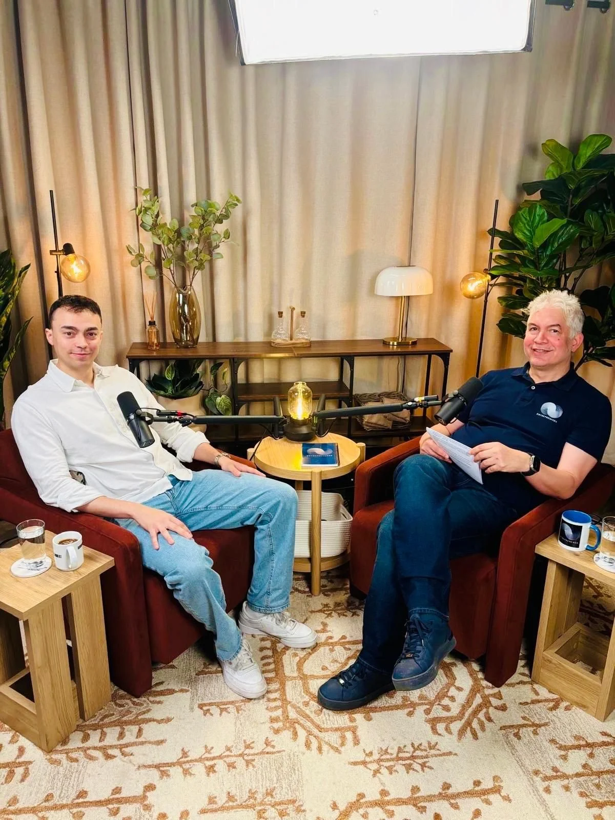 Two men sit in armchairs in a living room, with microphones and recording equipment, engaged in a podcast or interview, surrounded by plants and warm lighting.