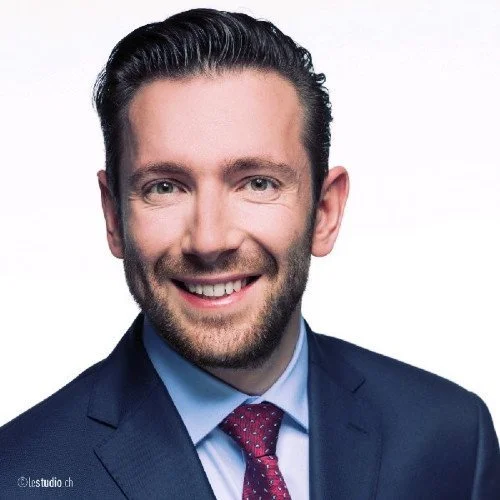 A close-up professional headshot of a man with short dark hair, a beard, and blue eyes, wearing a dark blue suit, light blue shirt, and maroon tie, smiling against a white background.