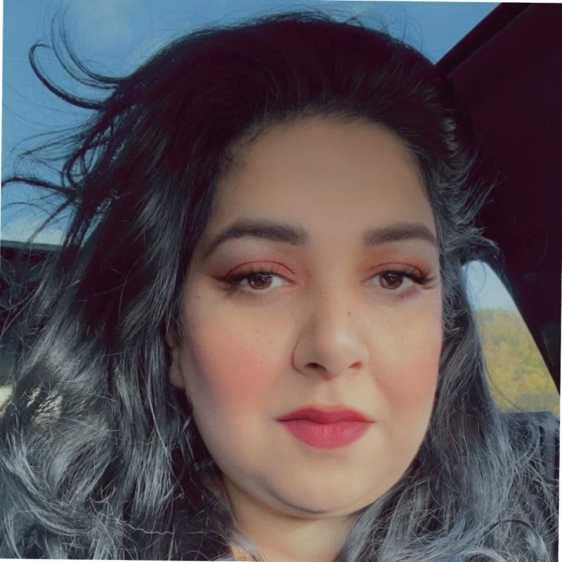 Close-up selfie of a woman with dark and gray wavy hair, wearing makeup with pink eyeshadow and lipstick, inside a vehicle.