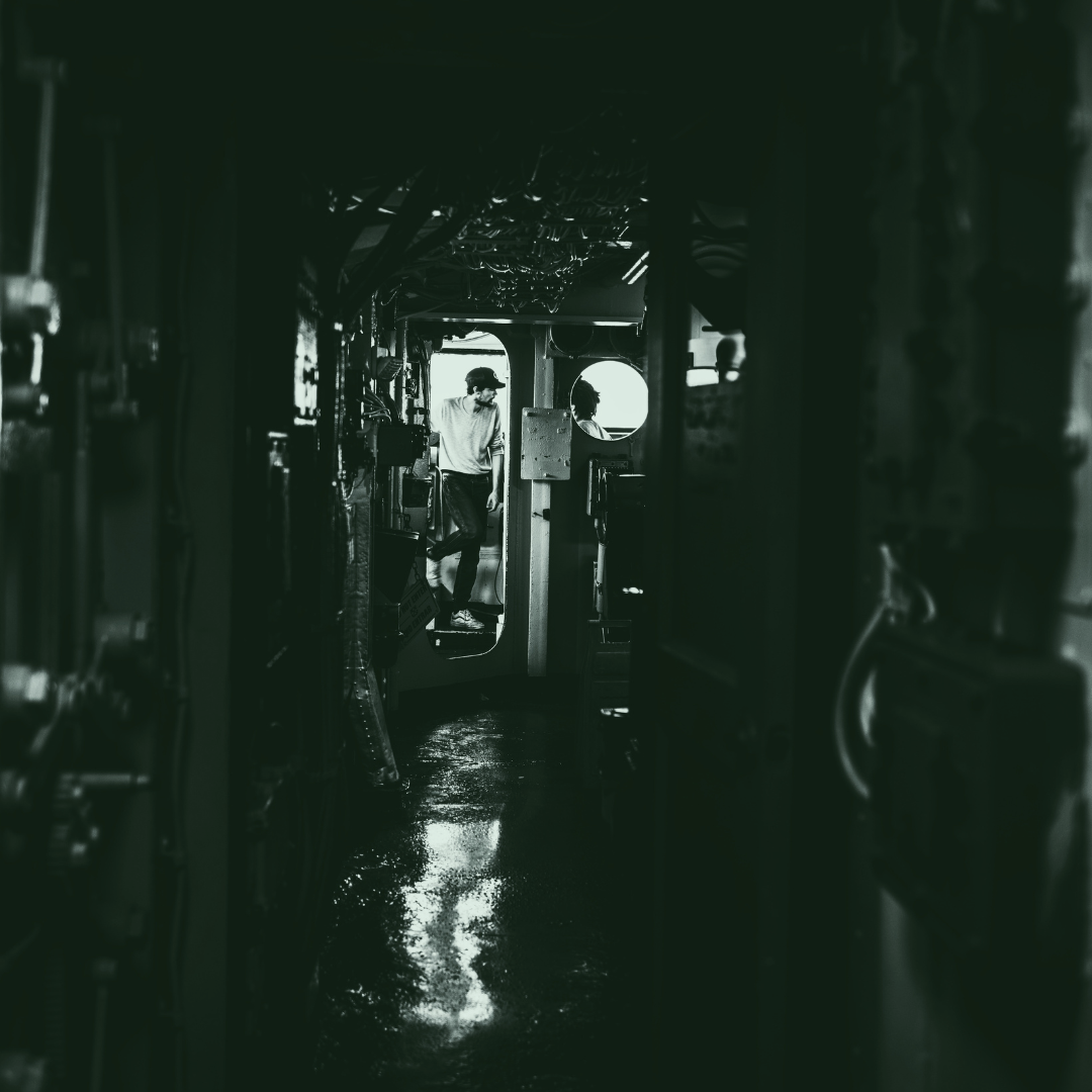 A black and white photo of a man standing inside a narrow, industrial corridor, with various machinery and pipes around him, looking out through a circular window.