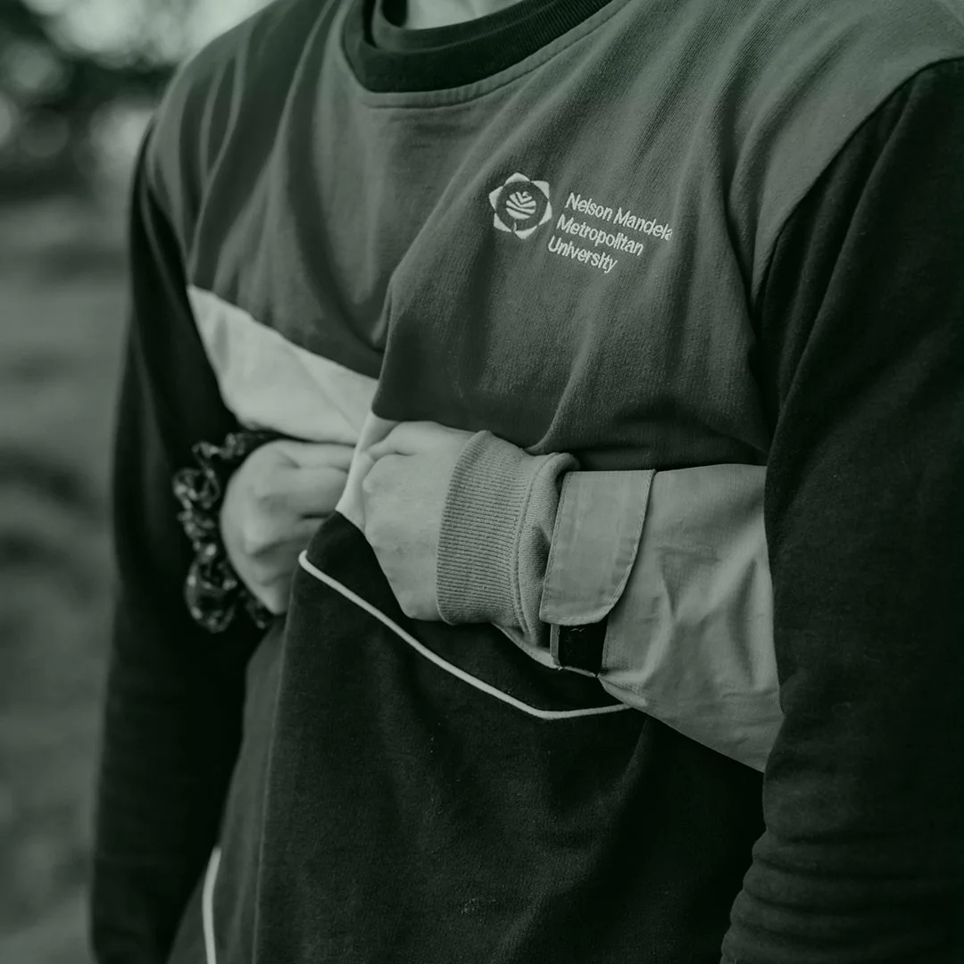 Close-up of a person wearing a Nelson Mandela Metropolitan University sweatshirt with an armband, holding another person's arm in a supportive gesture, outdoors.