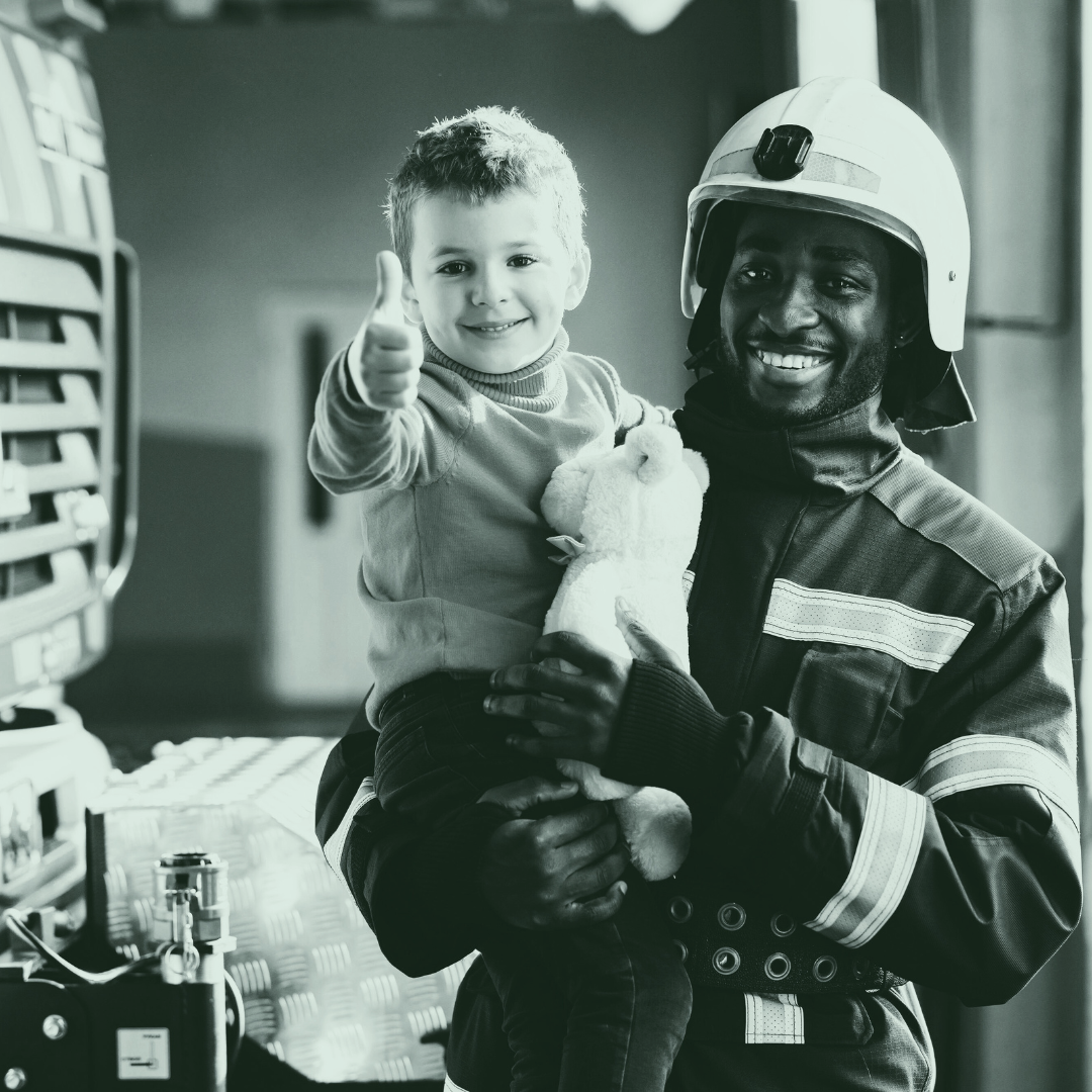 A firefighter holding a young boy, who is giving a thumbs-up gesture, in a fire station. The boy holds a stuffed animal and is smiling. The firefighter is wearing a helmet and protective gear, both smiling.