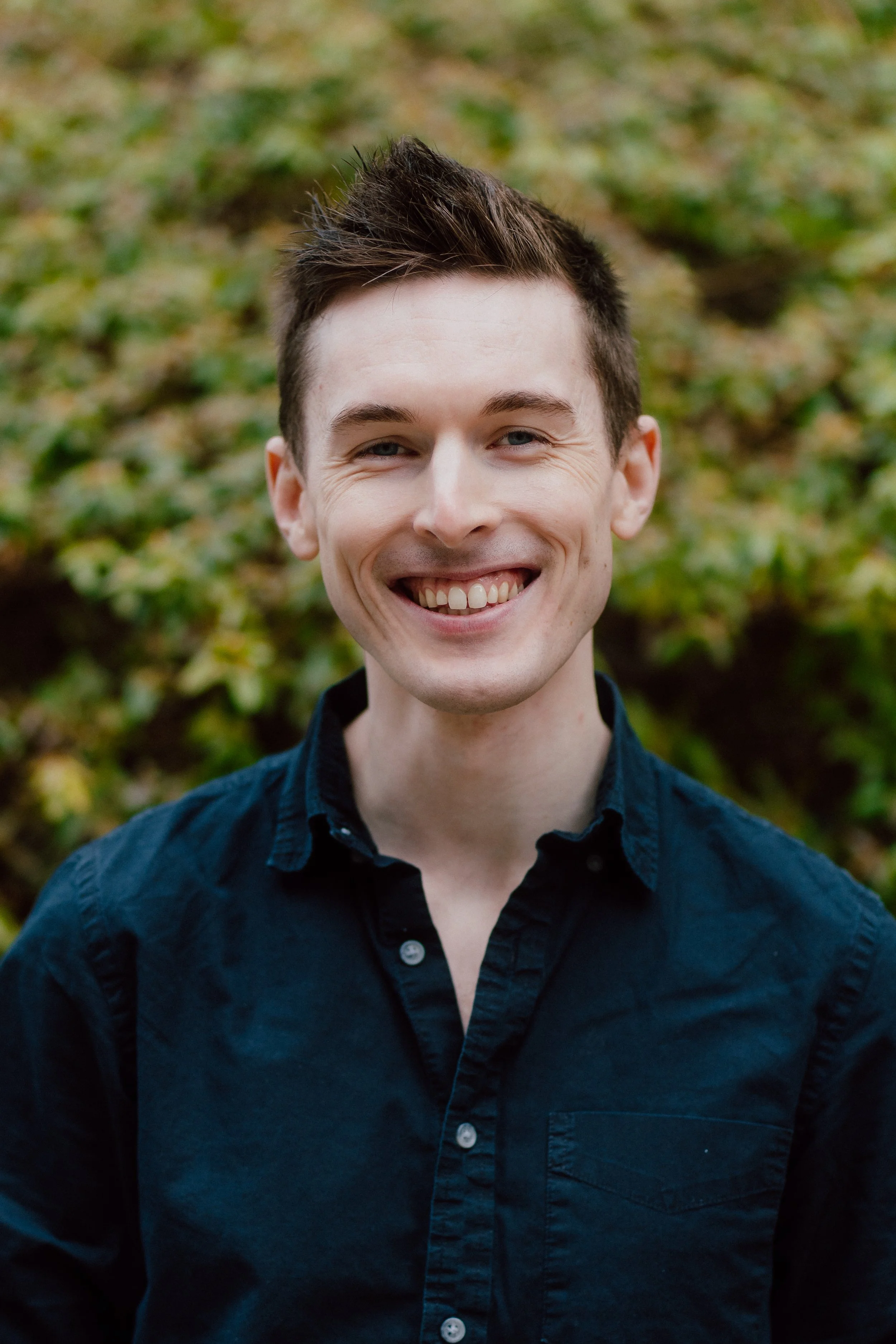 Smiling young man with short brown hair wearing a dark button-up shirt outdoors, with blurred green foliage in the background.