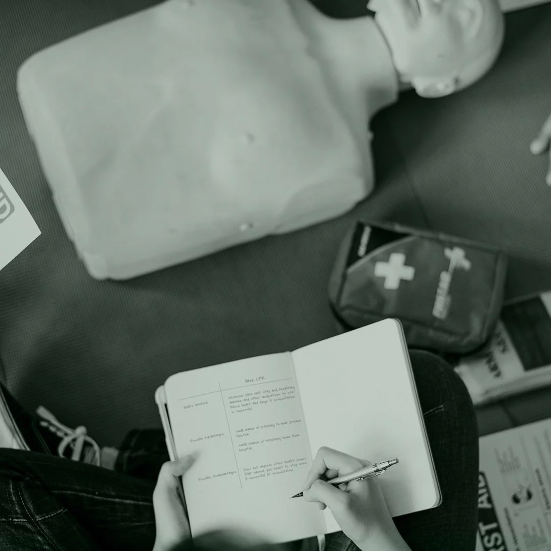 Person sitting and writing in a notebook, with a medical first aid kit and a large container on the seat next to them.