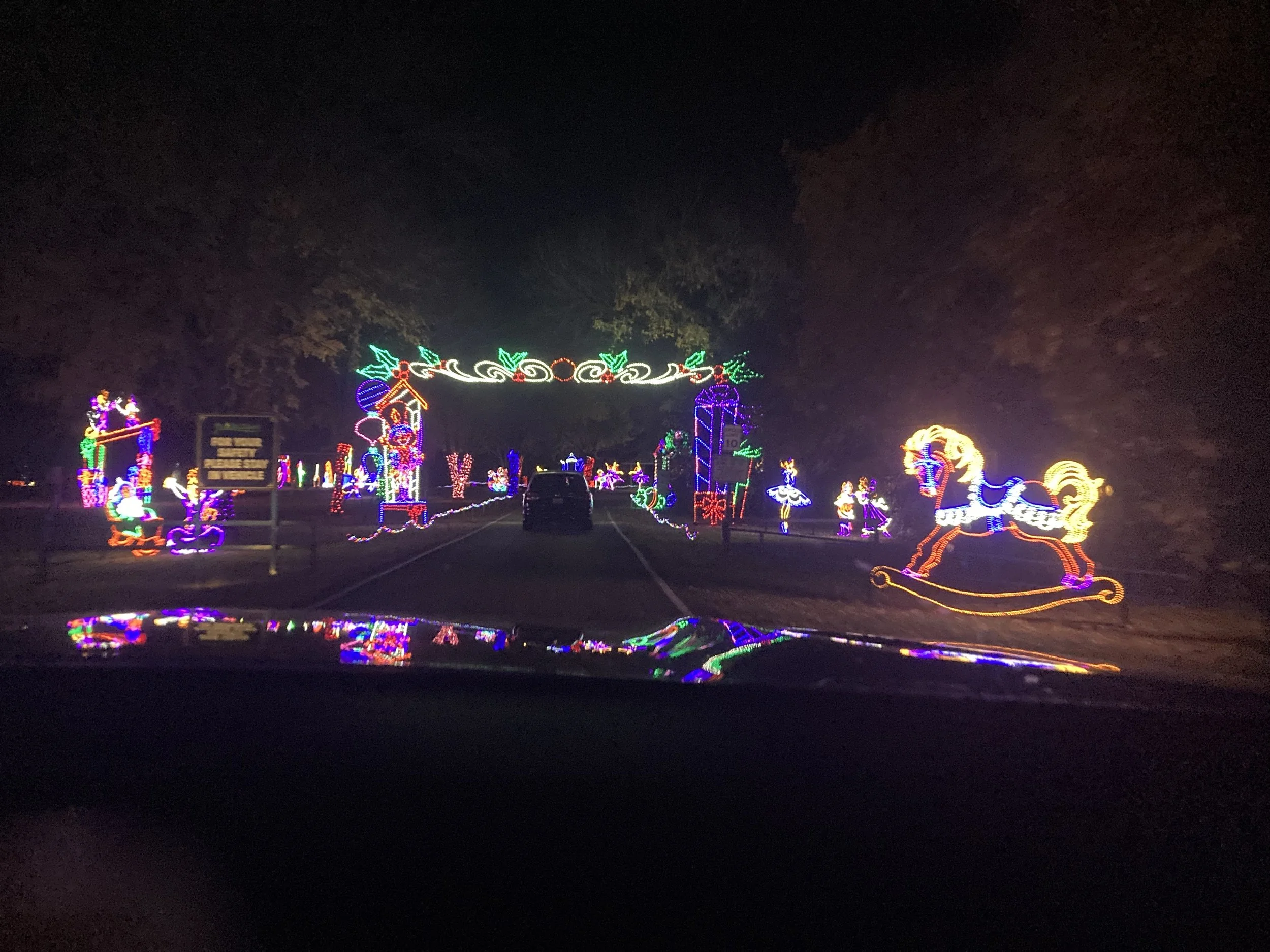 Colorful Christmas light display on a night street featuring lit figures of Santa Claus, reindeer, a carousel horse, presents, and holly, viewed from inside a vehicle.