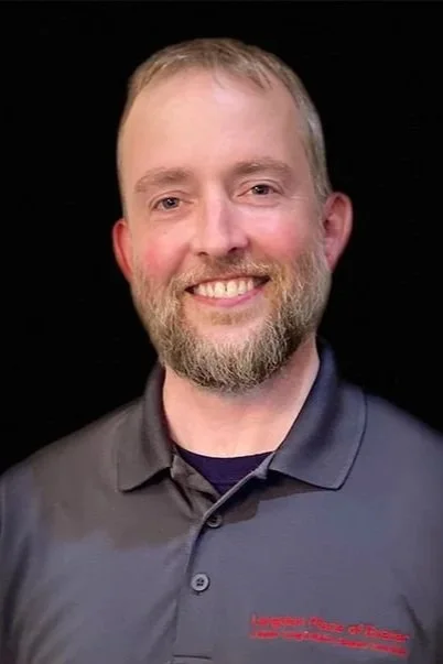 A smiling man with a beard and short hair, wearing a dark gray polo shirt with red and white text, against a black background.