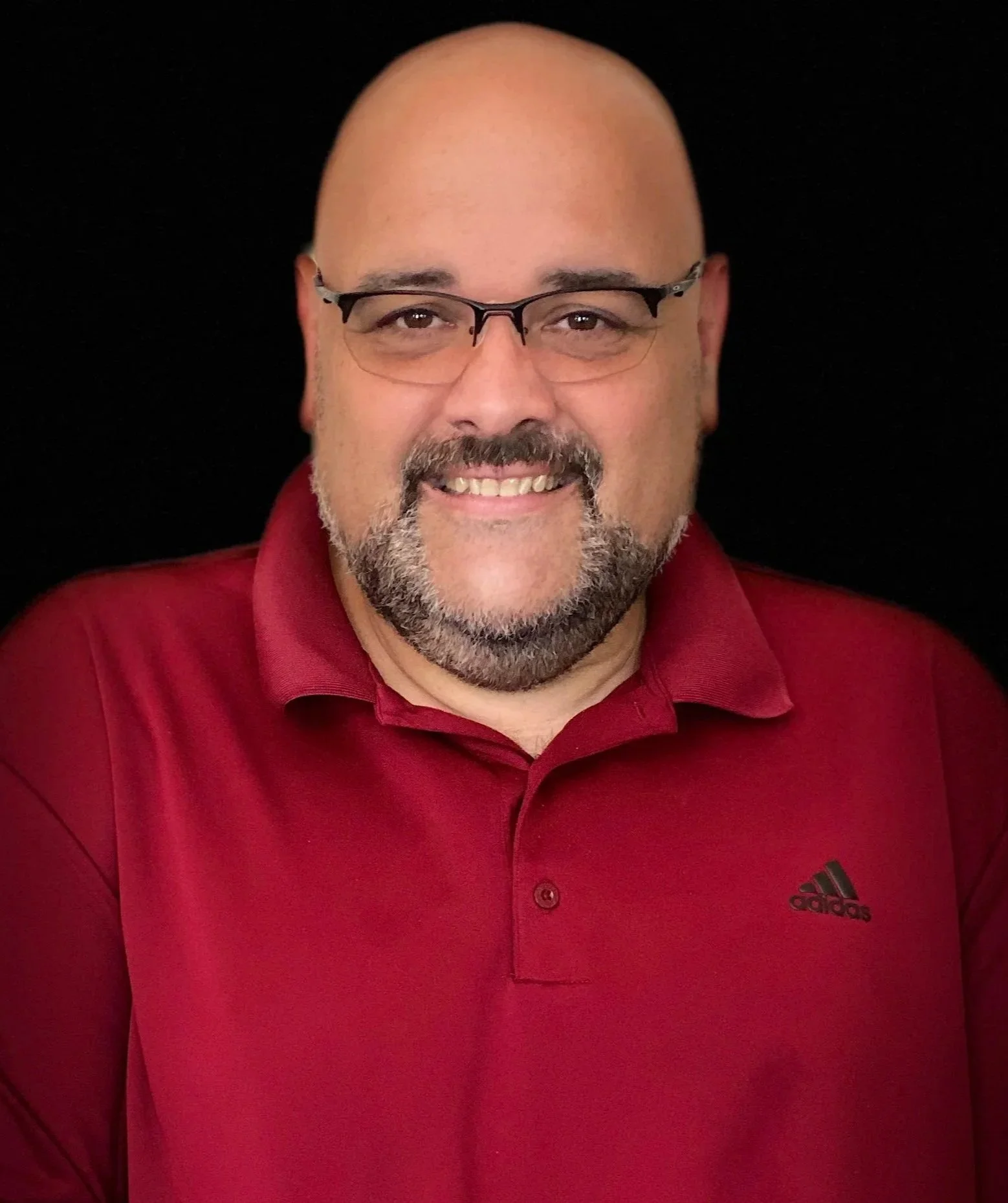 A man with glasses, a beard, and a bald head, smiling, wearing a red Adidas polo shirt against a black background.