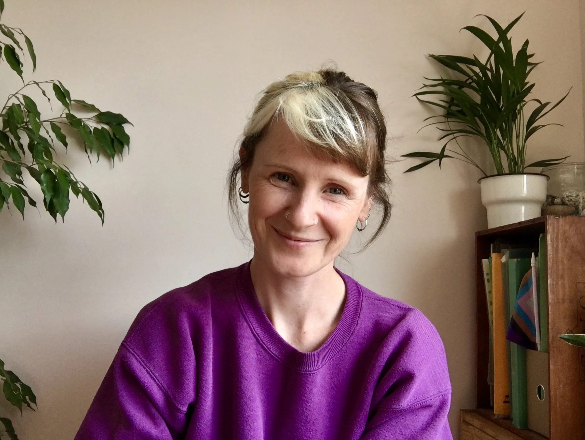 Smiling white woman wearing a purple jumper, with brown hair and blonde streak, seated in front of plants and books.