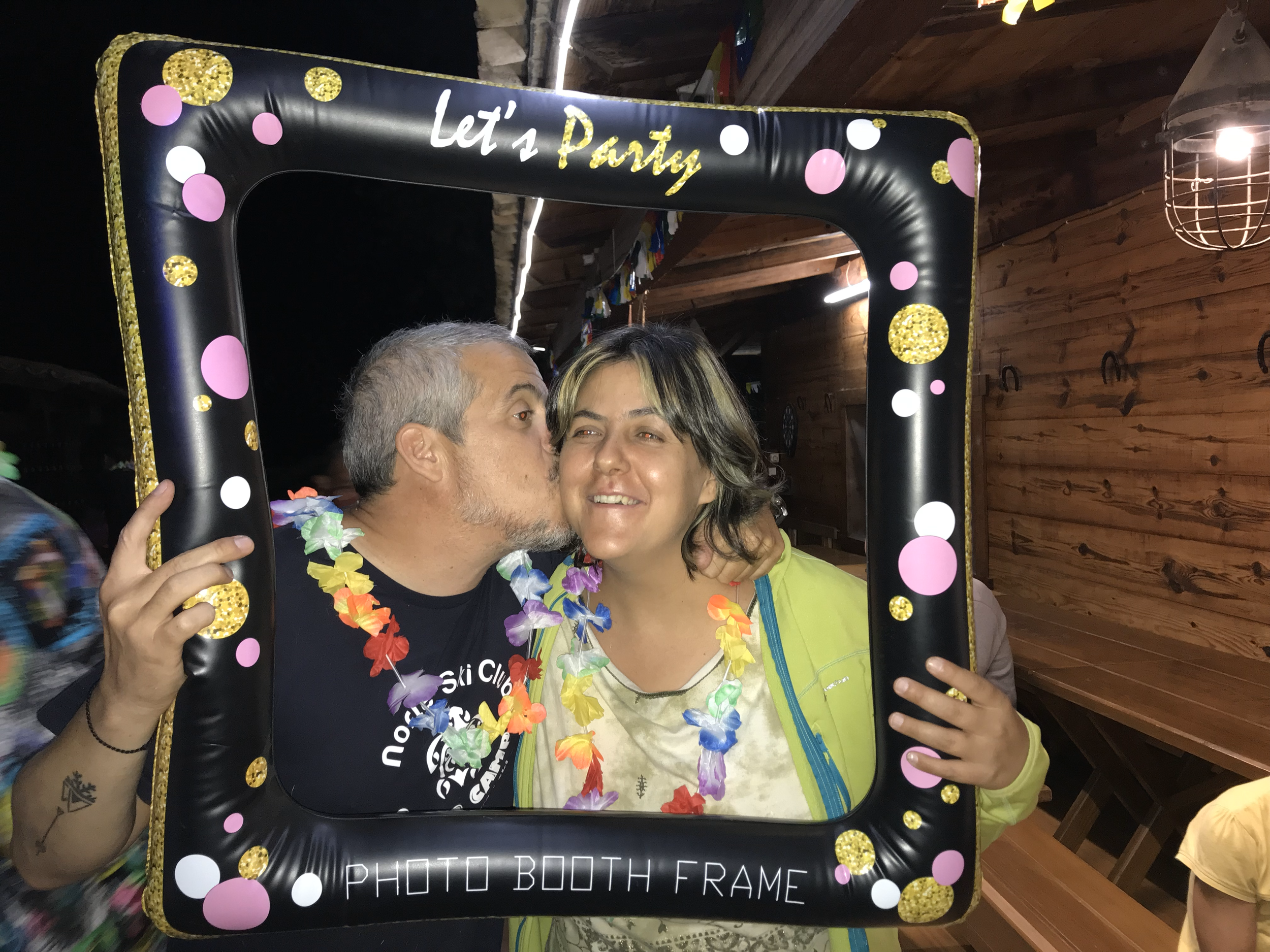 Dos personas sonríen dentro de un marco inflable decorado con colores brillantes y la frase "Let's Party" durante una fiesta. Una persona le da un beso en la mejilla a la otra, ambos llevan collares de flores.