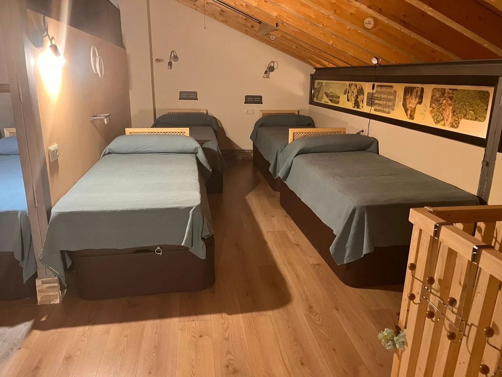 Cuatro camas individuales en una habitación con paredes blancas, techos de madera y suelo de madera. Cada cama tiene sábanas grises y cabeceras de madera. Hay lámparas de pared sobre las camas y un mural largo en la pared del fondo.