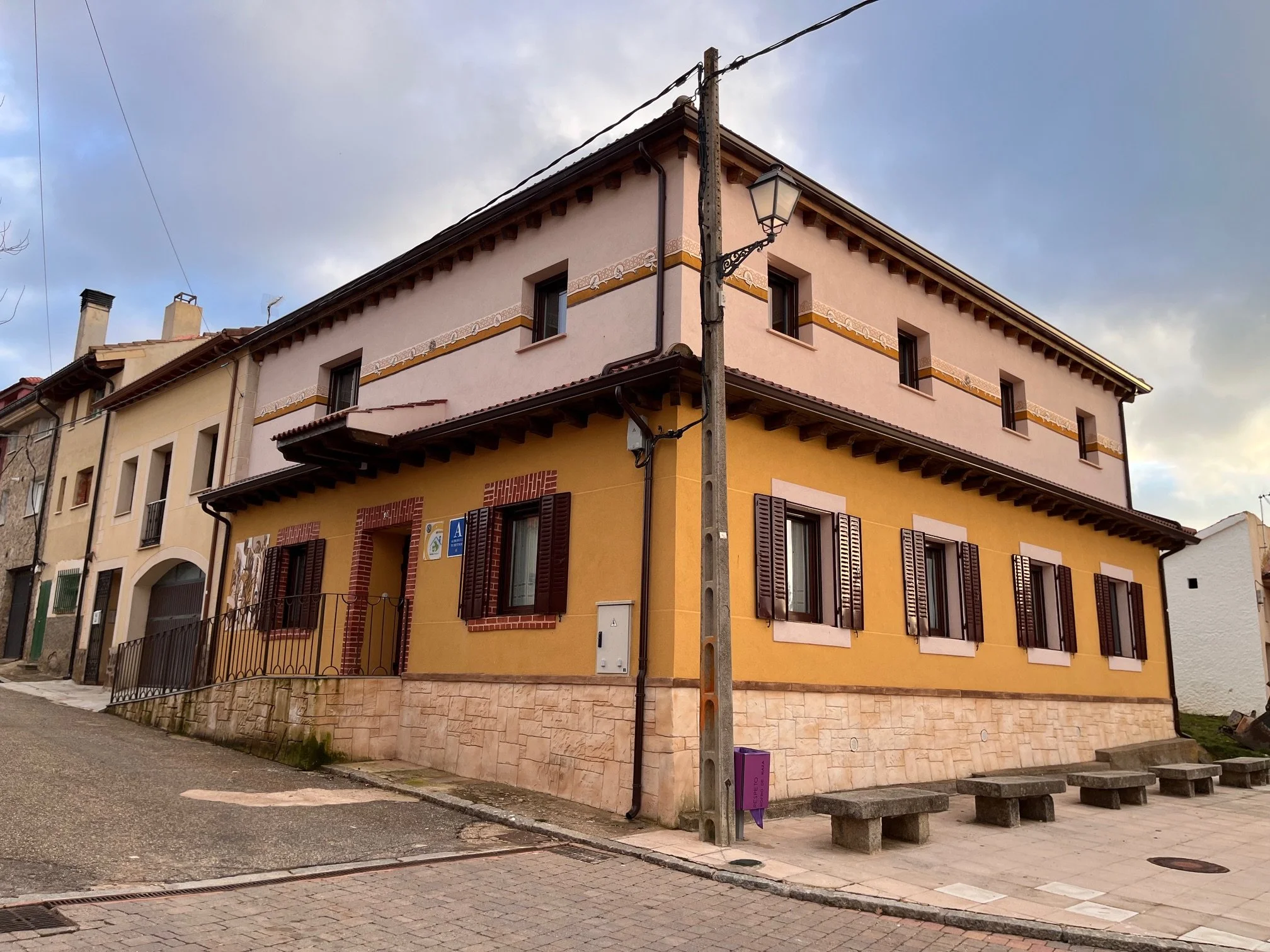 Edificio de dos pisos pintado en colores amarillo y rosa con ventanas con persianas de madera oscuras y una lámpara en un poste de luz en la esquina de la calle.