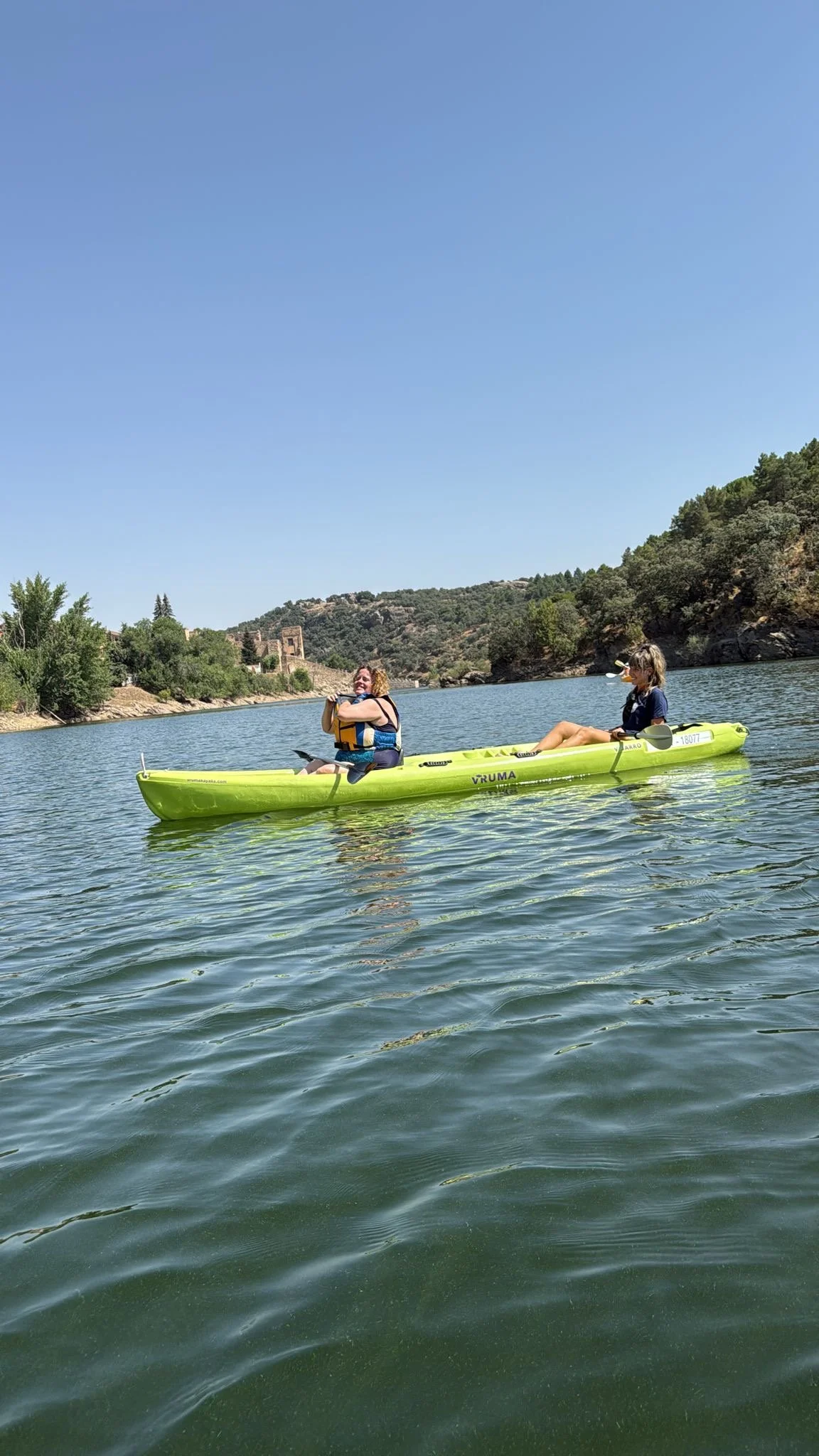 Dos personas remando en una kayak en un lago, rodeado de árboles y colinas con un cielo despejado.