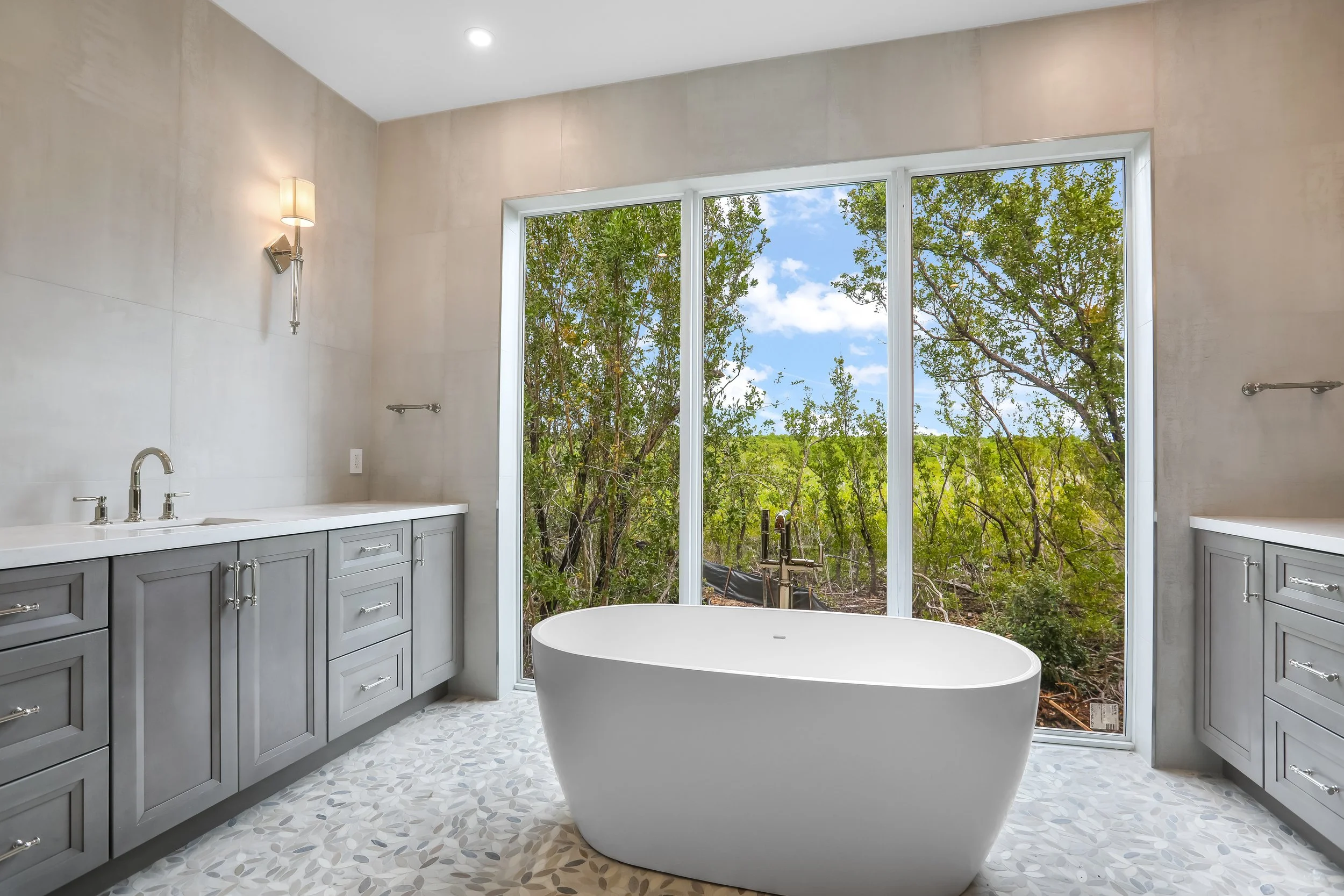 Luxury bathroom in a Key Largo, Florida home featuring a freestanding soaking tub, double vanity, and floor-to-ceiling windows overlooking tropical greenery.