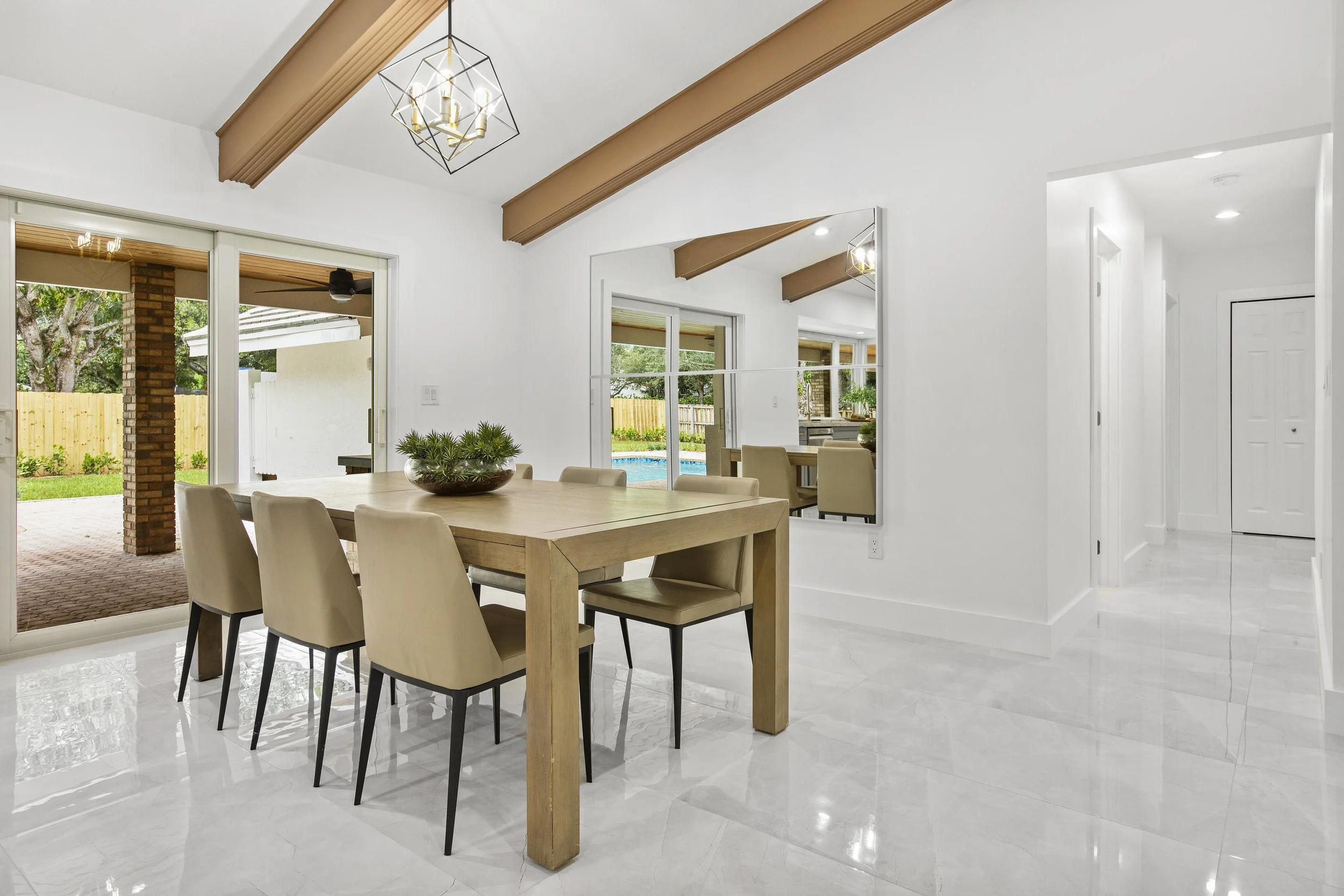 Interior of a bright, modern dining room with a wooden table, beige chairs, a decorative bowl of green plants, white walls, large sliding glass doors opening to a backyard with trees and a swimming pool, and a kitchen area visible in the background.
