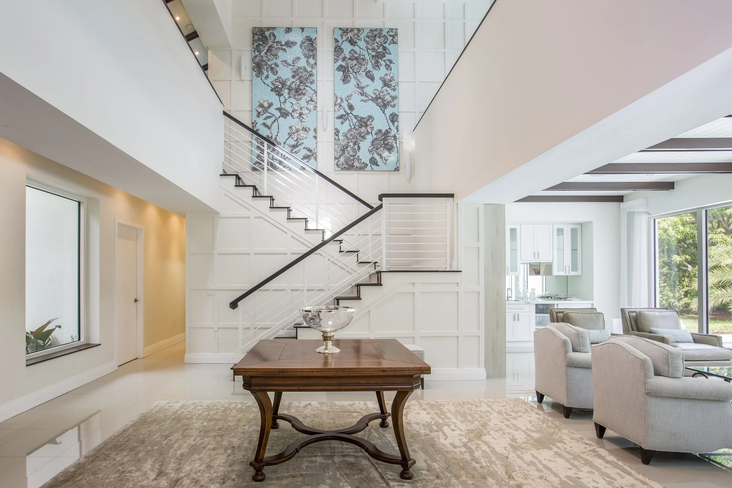 Bright and spacious living room with white walls and large windows, a staircase with black handrail, a wooden dining table, a beige area rug, glass bowl centerpiece, and a view of a green garden outside.