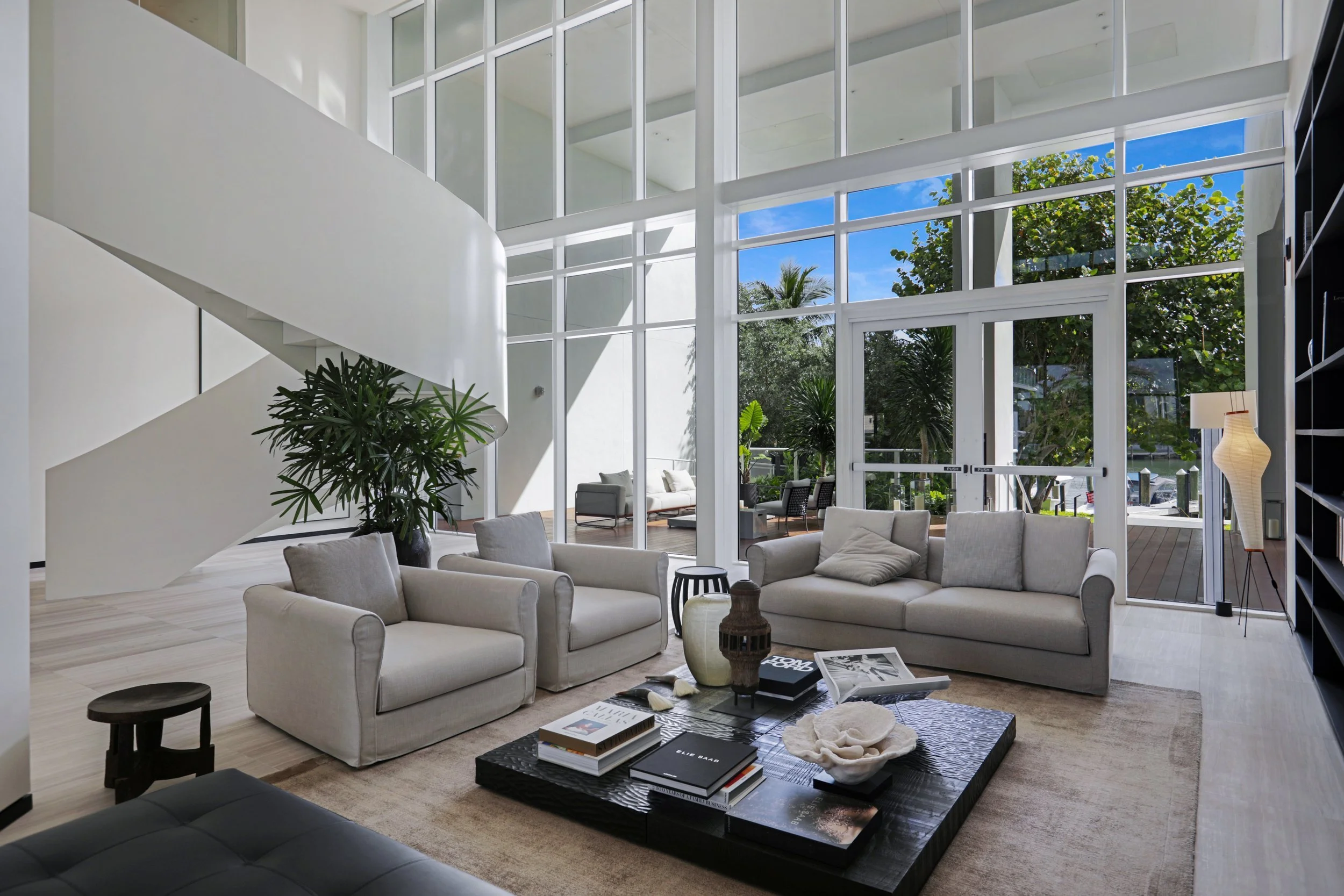 A spacious modern living room with large glass windows and doors, white sofas, a black coffee table with books and decorative items, a tall potted plant, and visible outdoor greenery and blue sky.